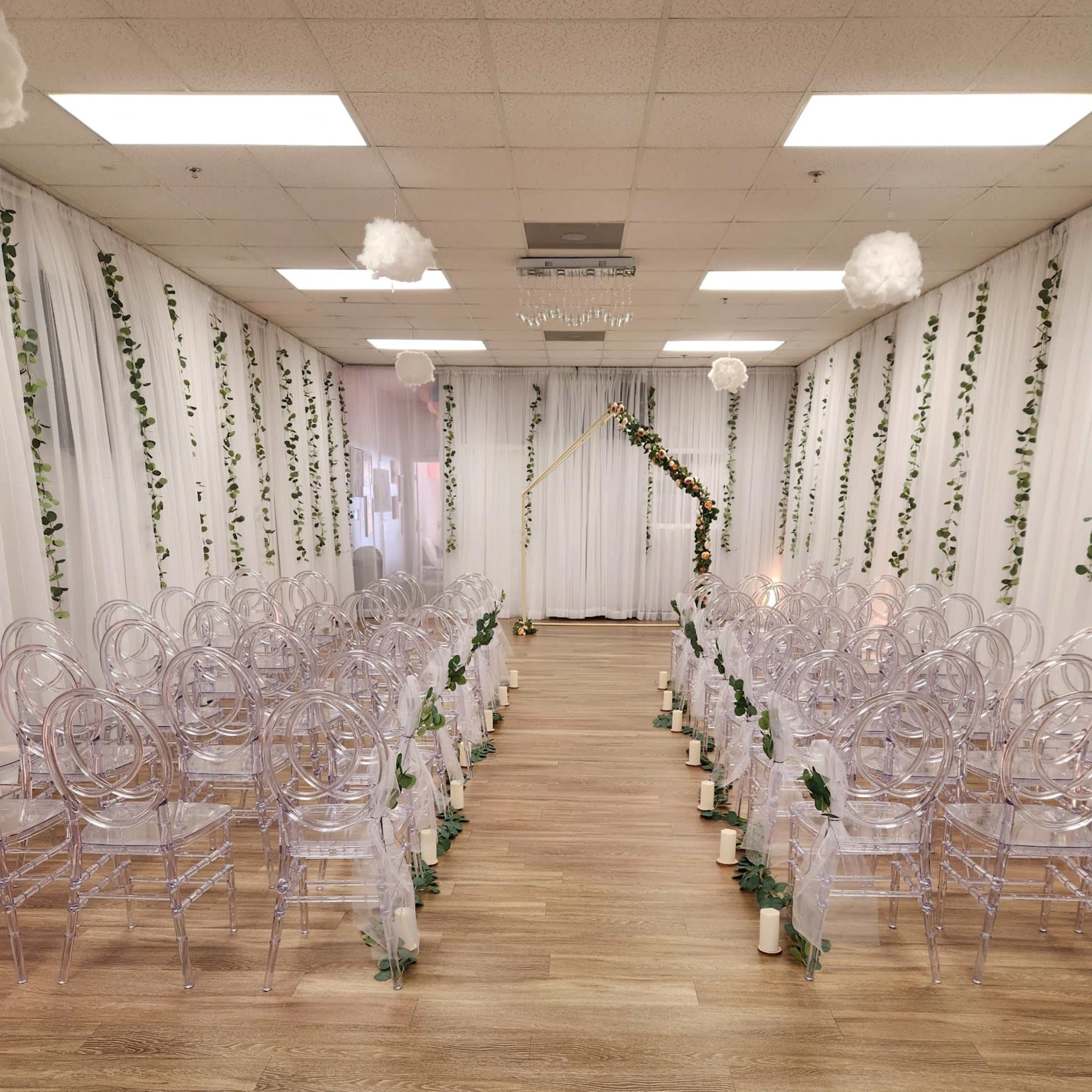 A wedding ceremony setup features clear acrylic chairs arranged in rows along a central aisle adorned with greenery and white fabric draping.
