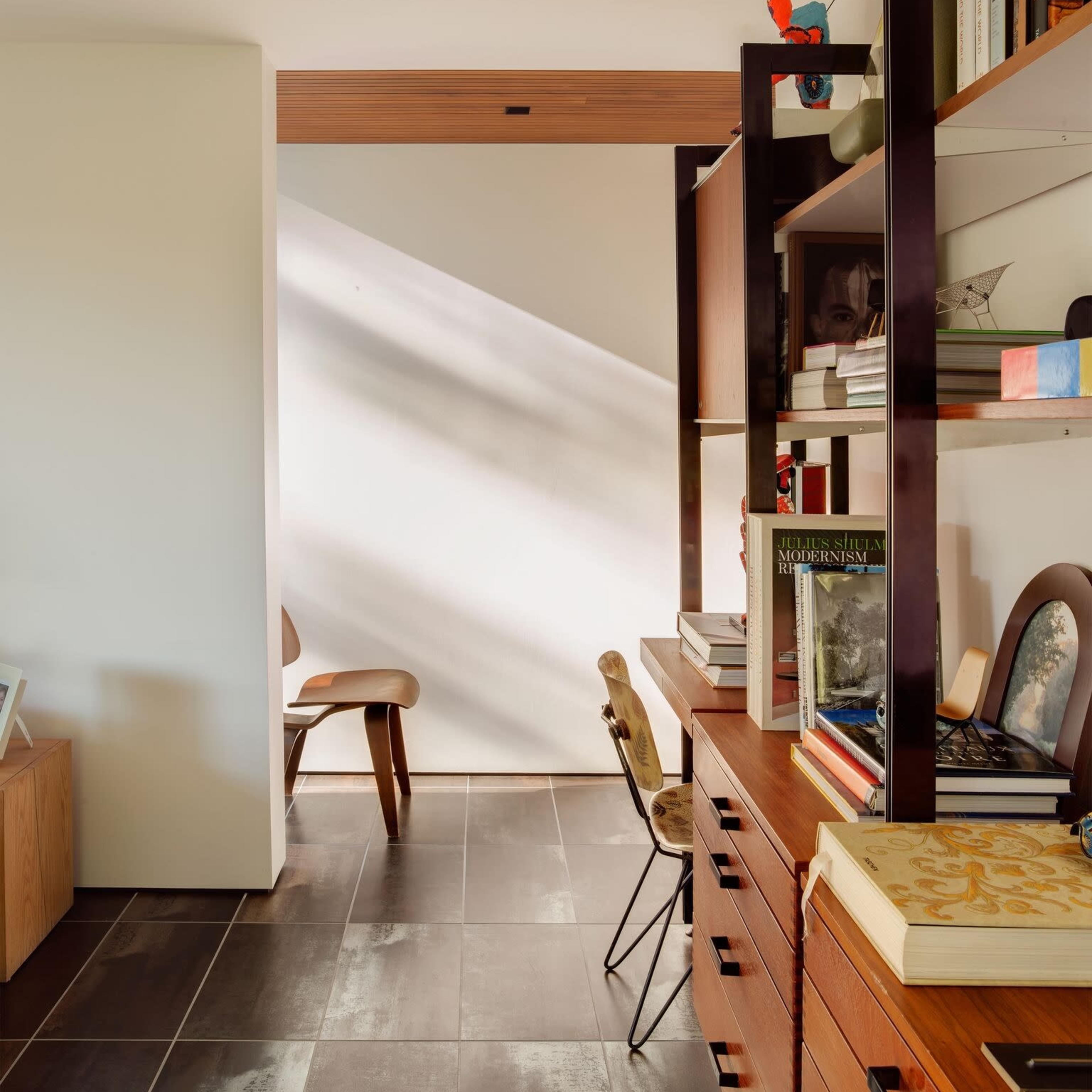 A modern, well-organized room features a bookshelf filled with books, a wooden desk with chairs, and natural light streaming through a door.