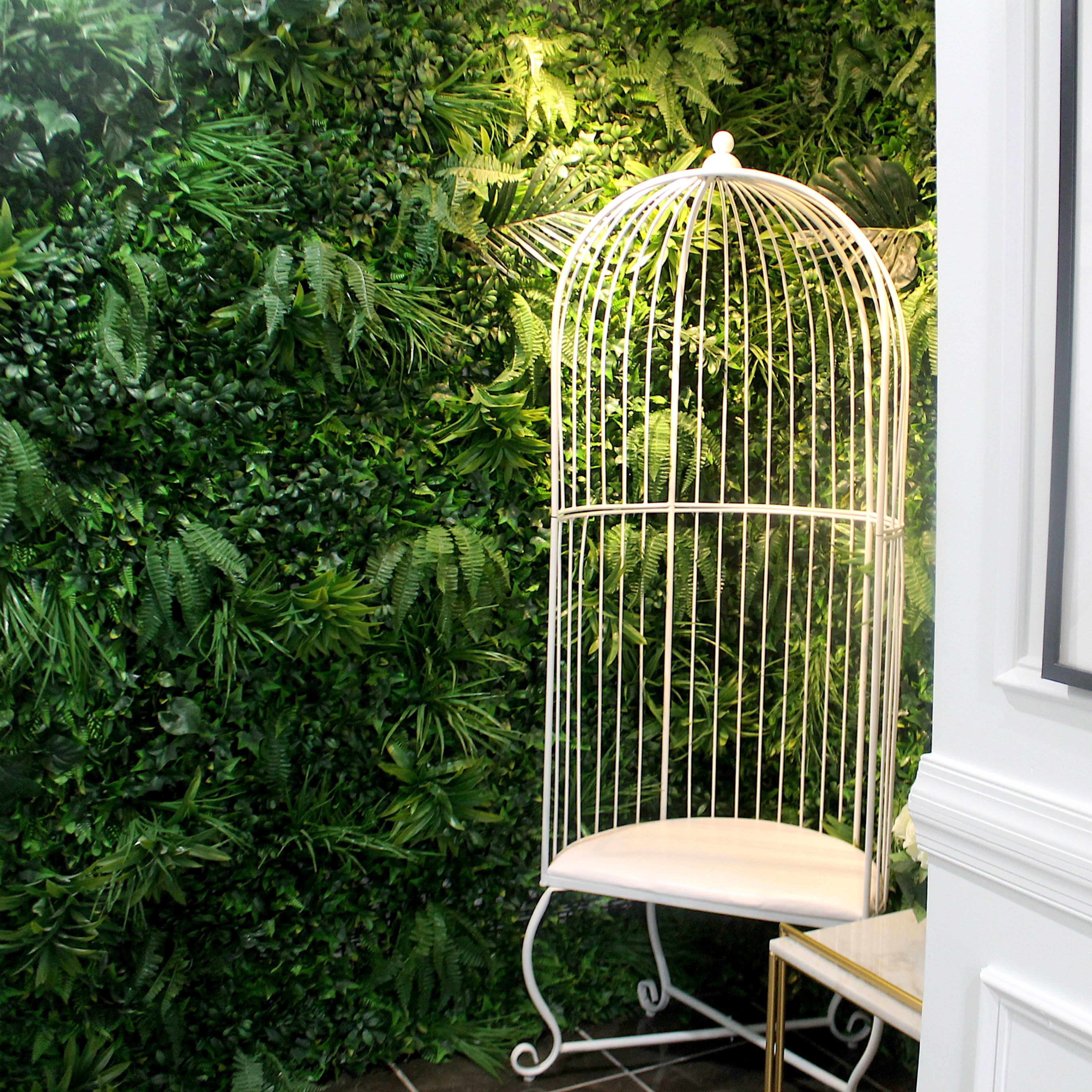 A white metal birdcage with a bench inside, positioned against a vibrant green wall of artificial foliage.