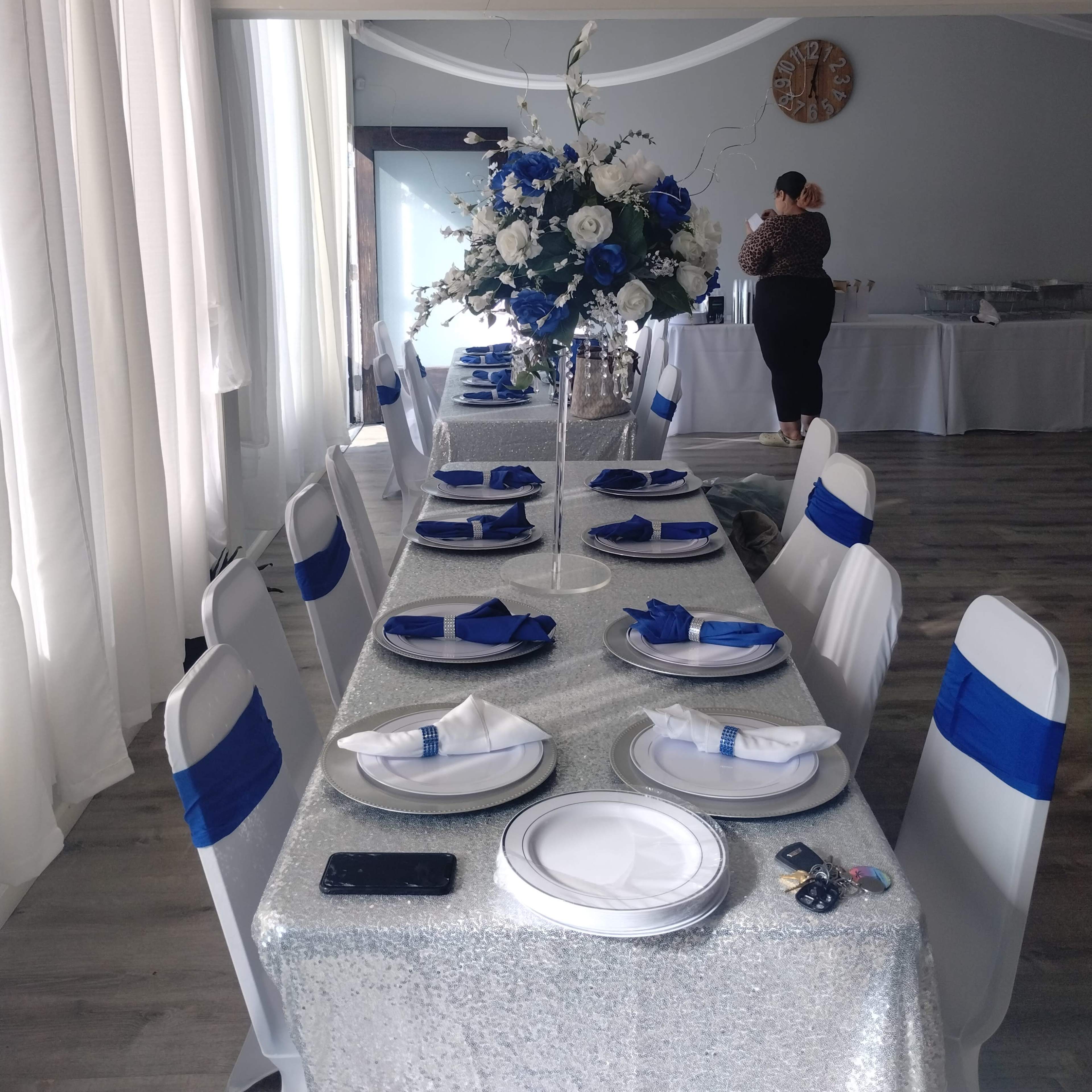 A long table is set with white plates and blue napkins, adorned with a floral centerpiece, in a room decorated with white drapes and a clock on the wall.