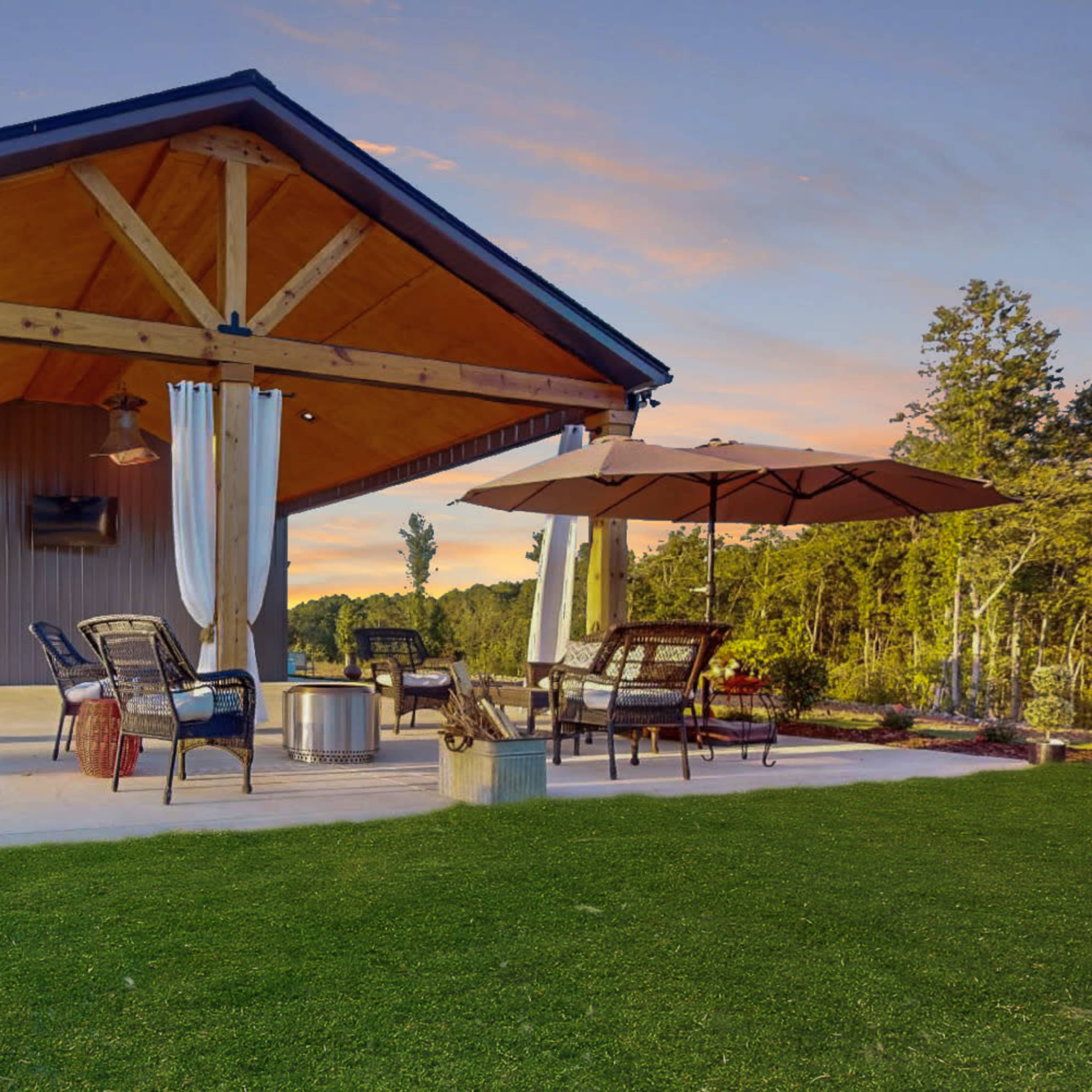 A modern outdoor pavilion with wooden beams, seating arrangements, and an umbrella, situated on a lawn beside trees under a sunset sky.