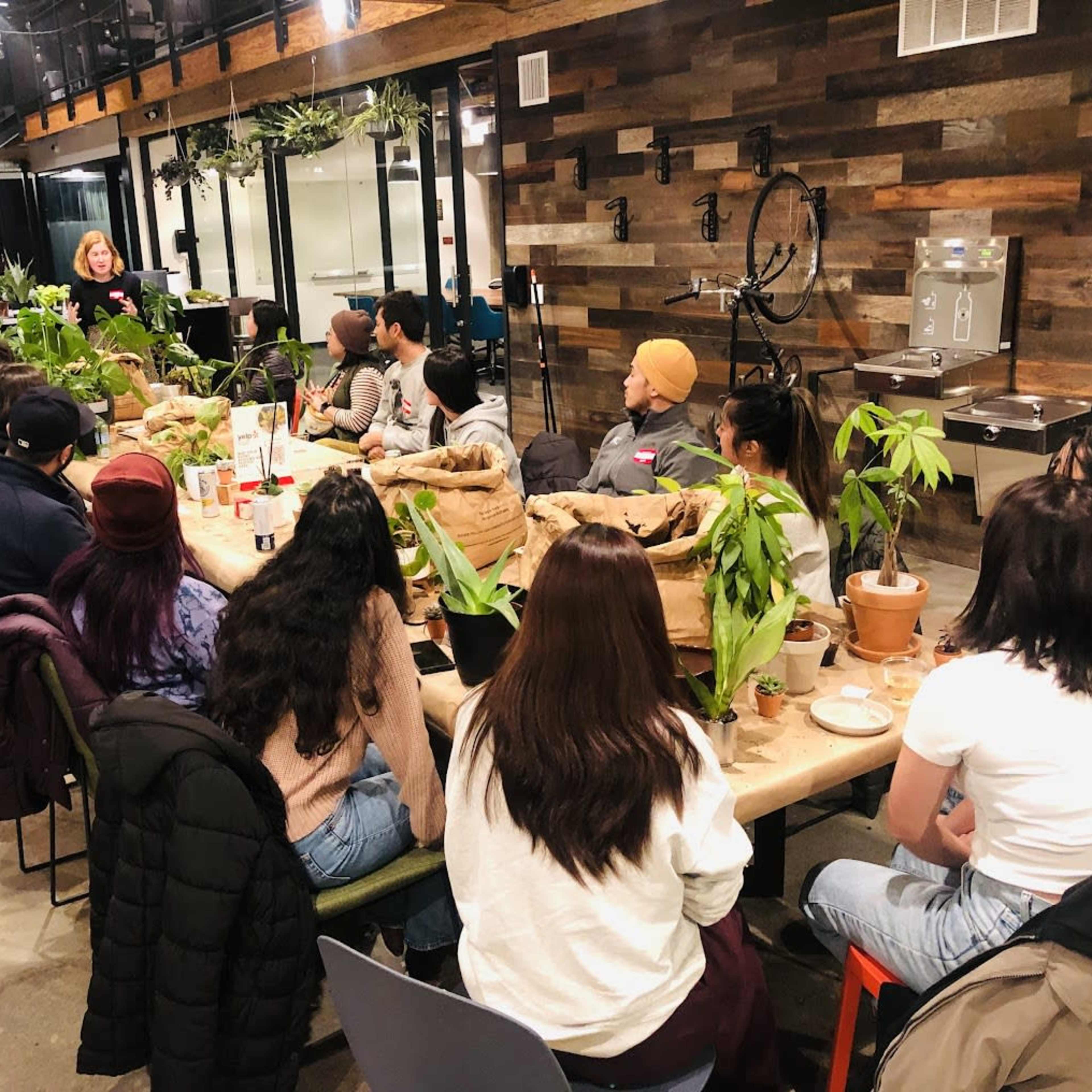 A group of people sits around a long table in a plant-filled space, listening to a speaker at the front.