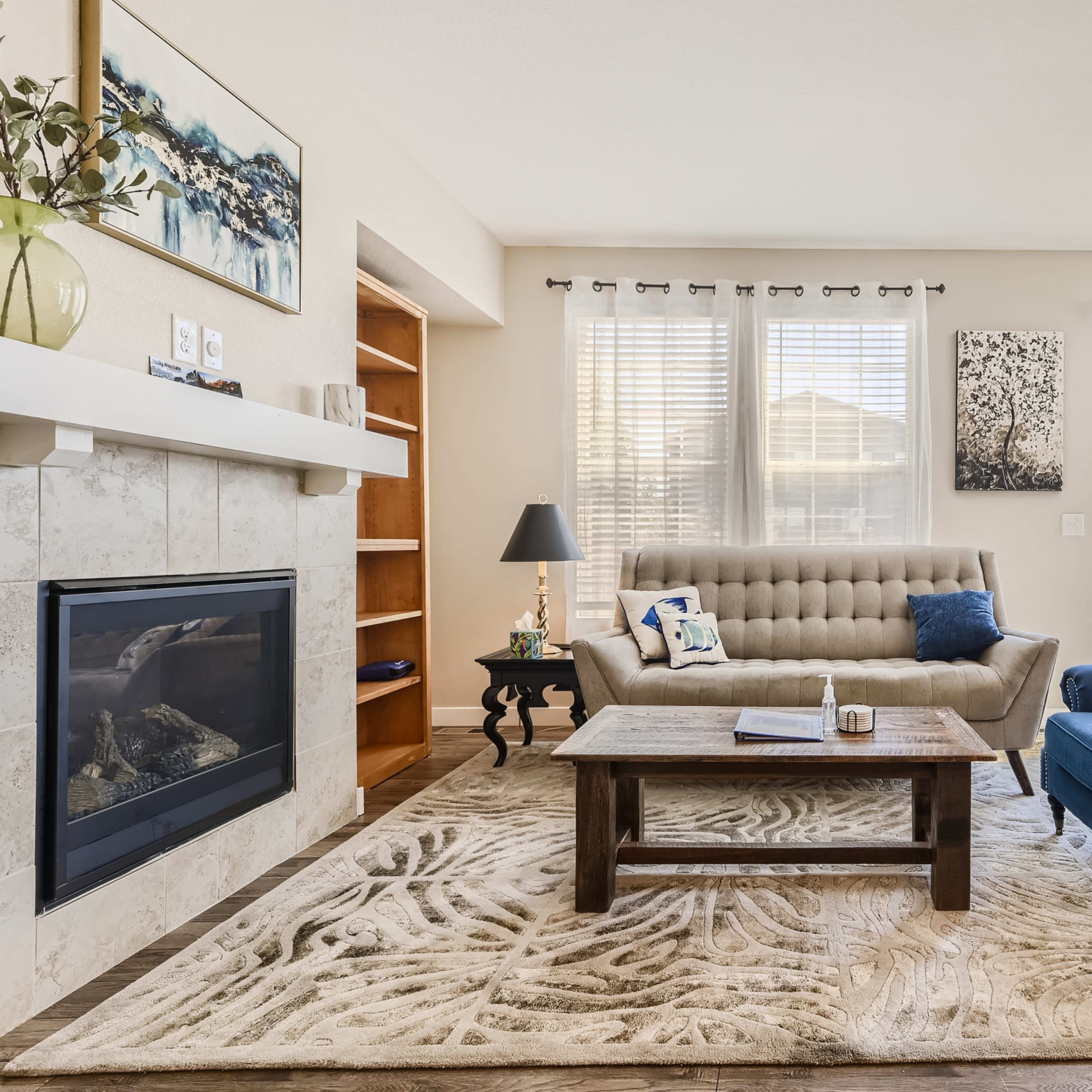 A living room with a beige sofa, a blue armchair, a coffee table on a patterned rug, and a fireplace, illuminated by natural light from large windows.