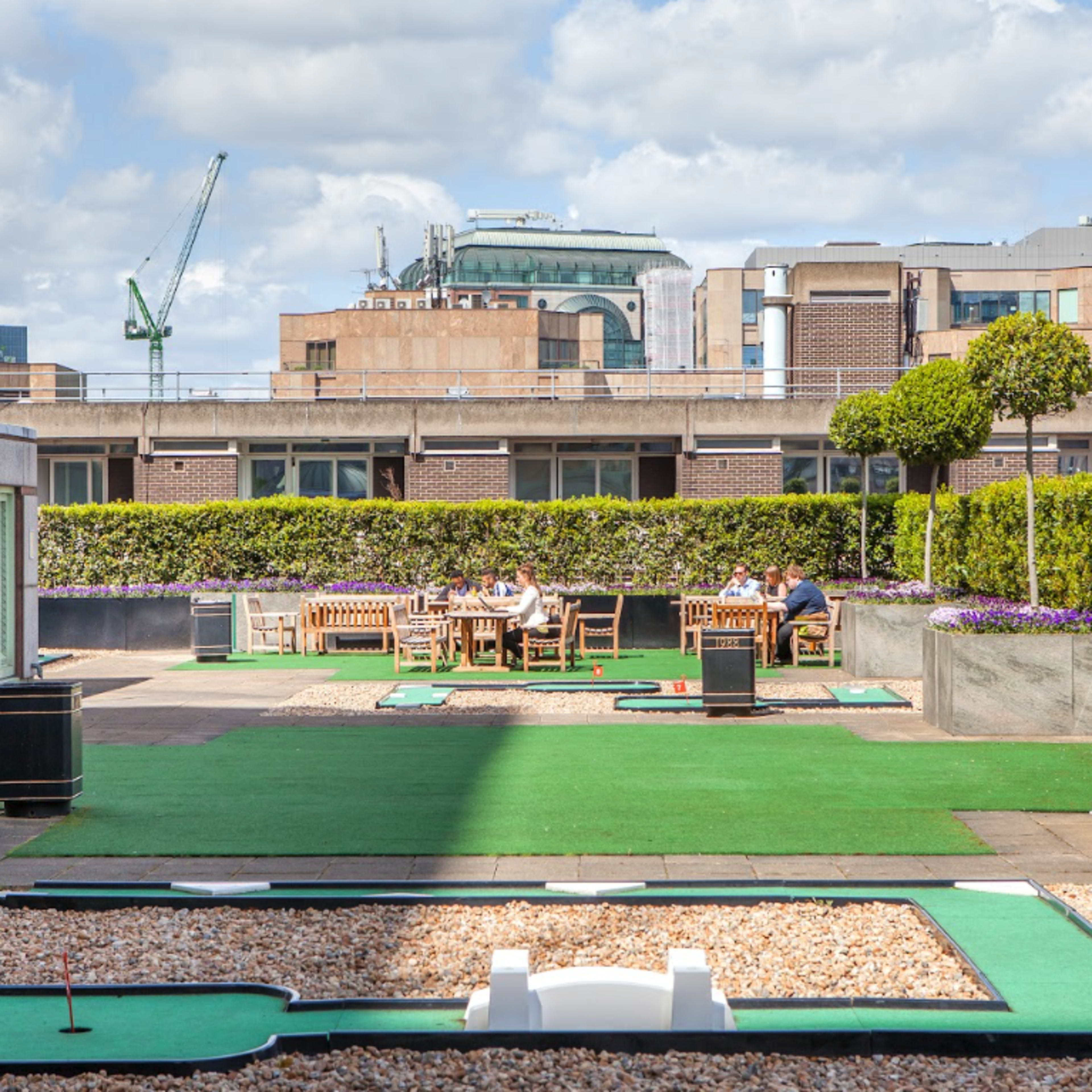 A rooftop miniature golf course features players enjoying the game among landscaped greenery and city buildings in the background.