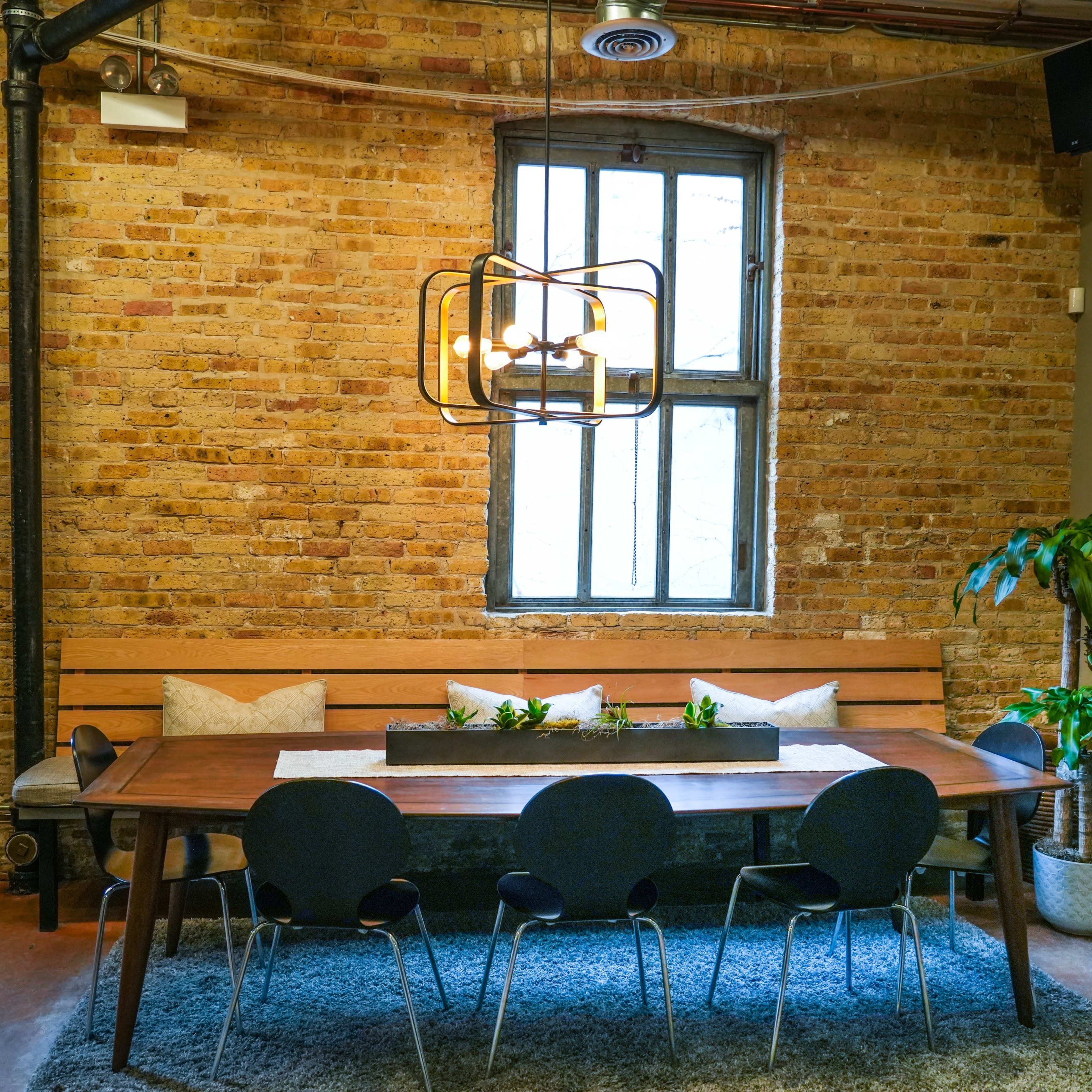 A wooden dining table with black chairs is set against a brick wall, featuring a bench along one side and plants beside the windows.