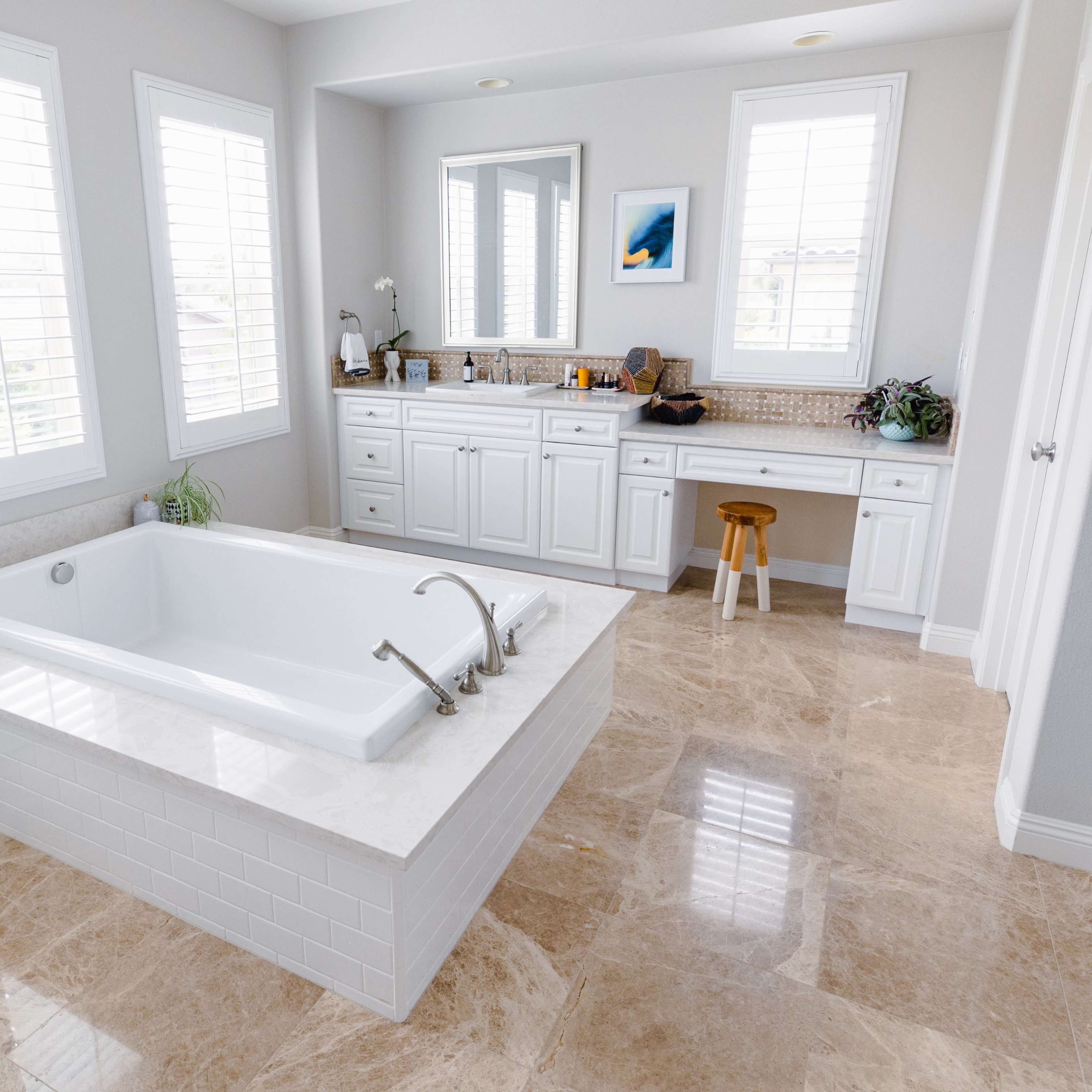 A modern bathroom featuring a freestanding bathtub on a marble platform, a double vanity with a mirror, and natural light filtering through white-shuttered windows.