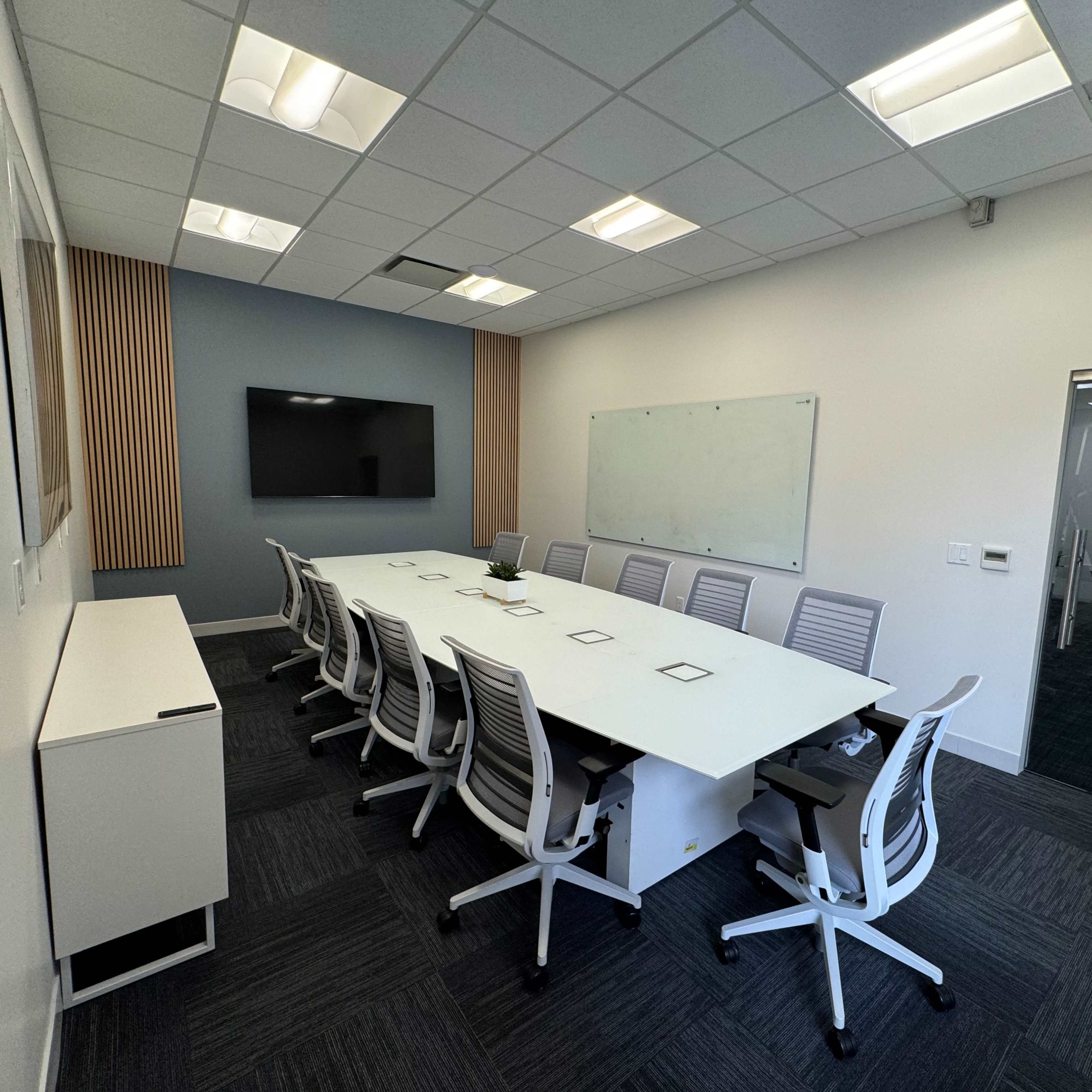 A modern conference room featuring a long white table surrounded by ergonomic chairs, a wall-mounted television, and a whiteboard.
