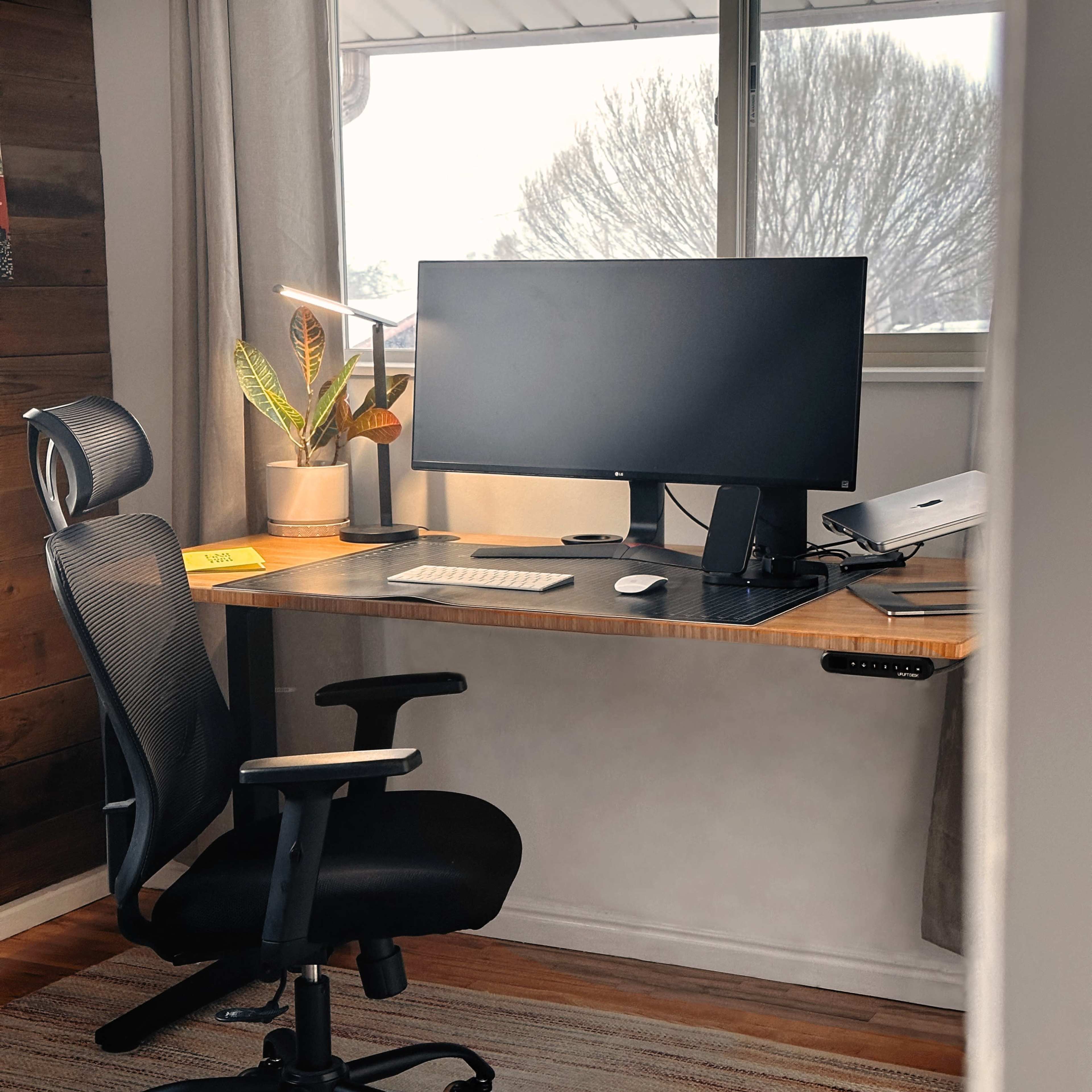 A modern workspace features a computer monitor, keyboard, and mouse on a wooden desk, accompanied by a rolling chair and a desk lamp, with a window offering a view of trees outside.
