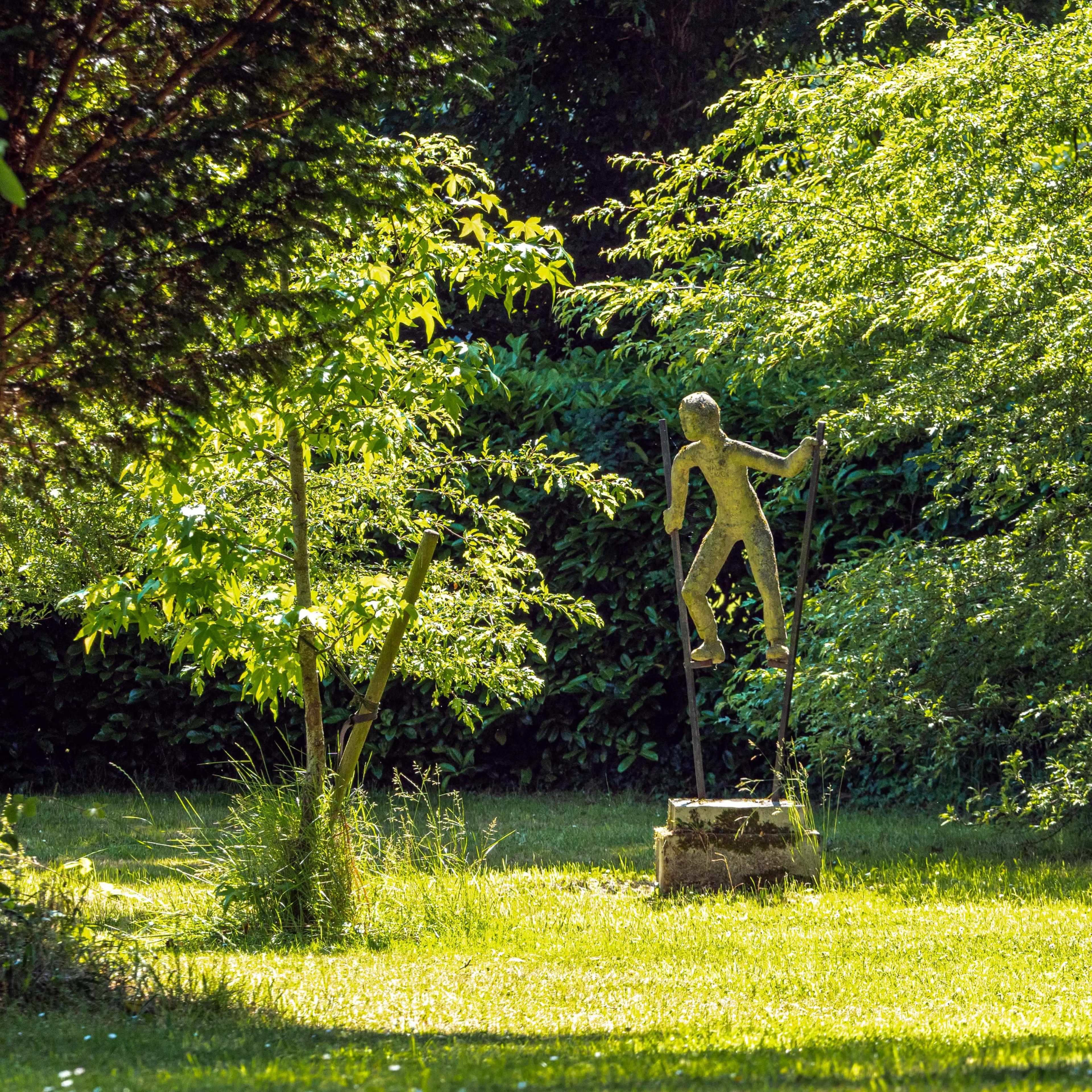 A metal sculpture of a figure climbing on a post stands in a lush green garden surrounded by trees.