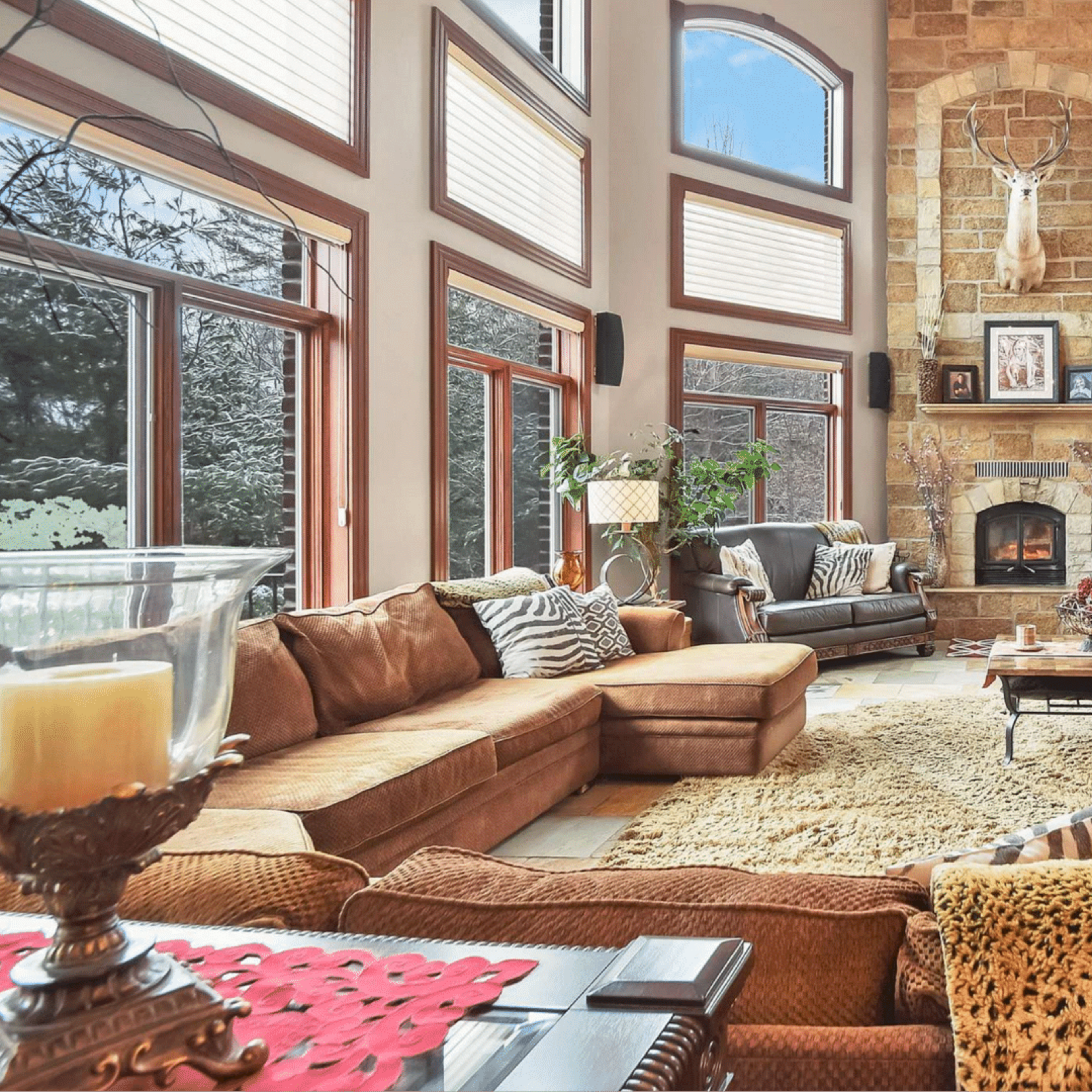 A spacious living room with a stone fireplace, large windows, and a combination of brown furniture and decorative elements.