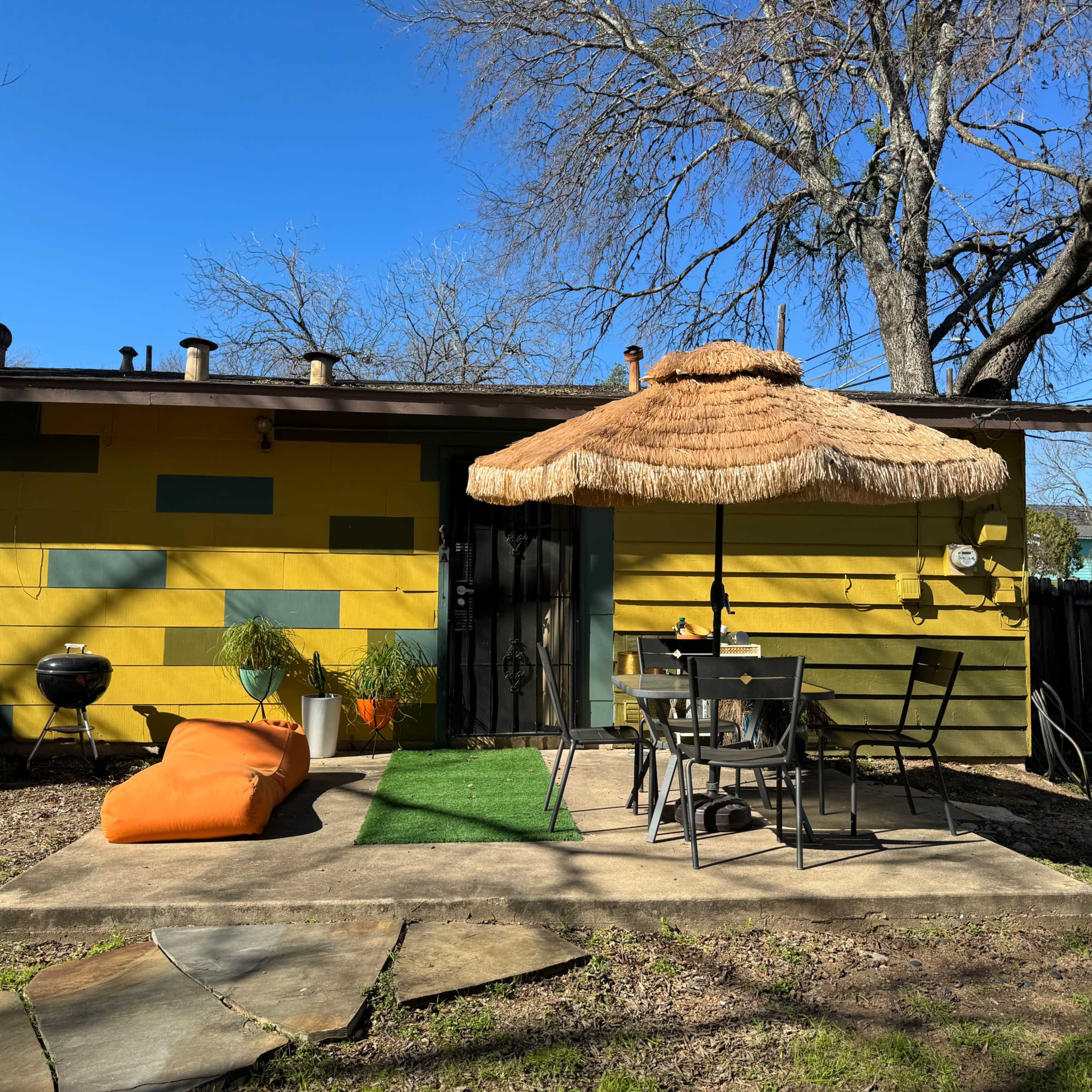 A backyard patio features a thatched umbrella over a table and chairs, with a bright yellow house and an orange bean bag nearby.