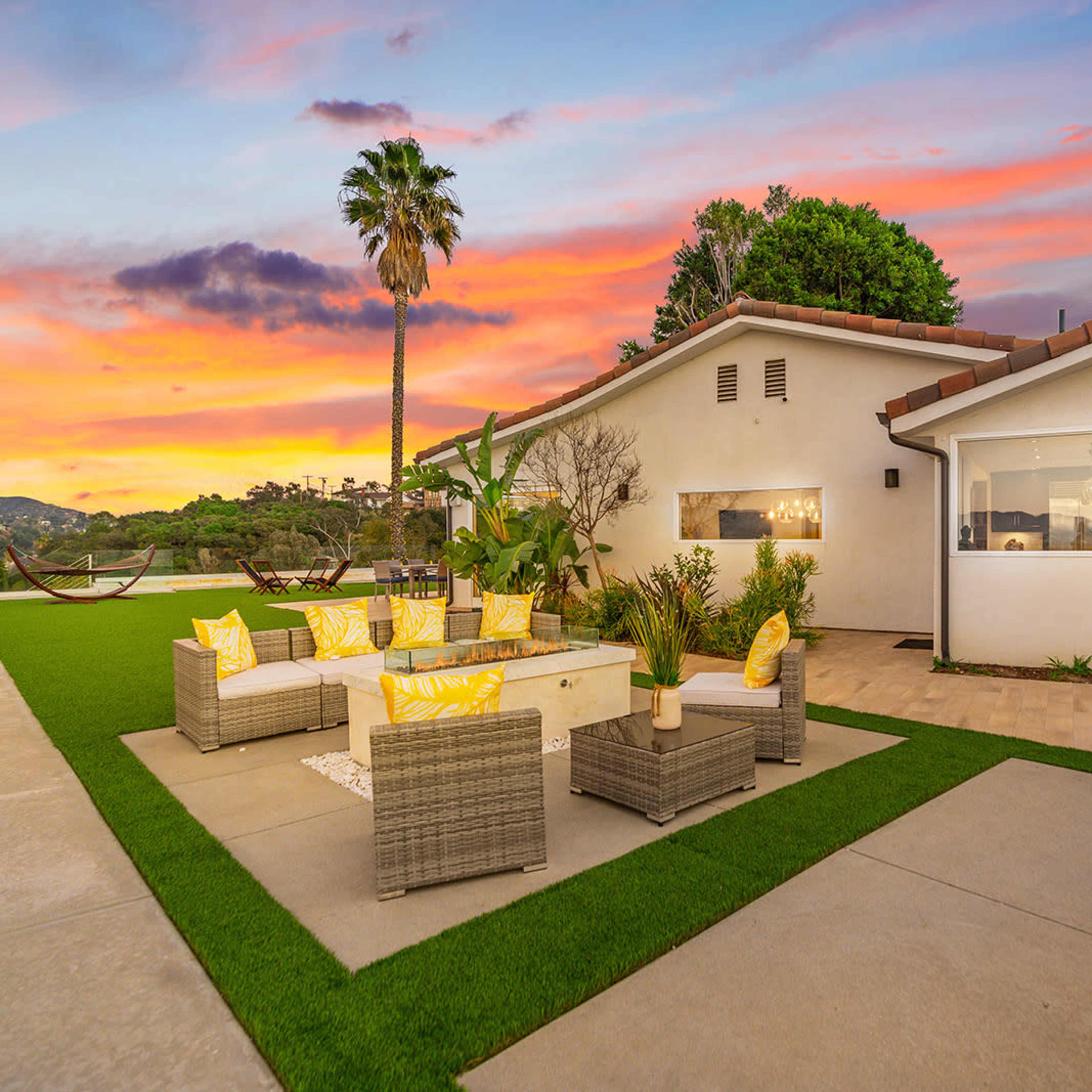 A modern outdoor seating area is set on a green lawn beside a house, with palm trees and a colorful sunset in the background.