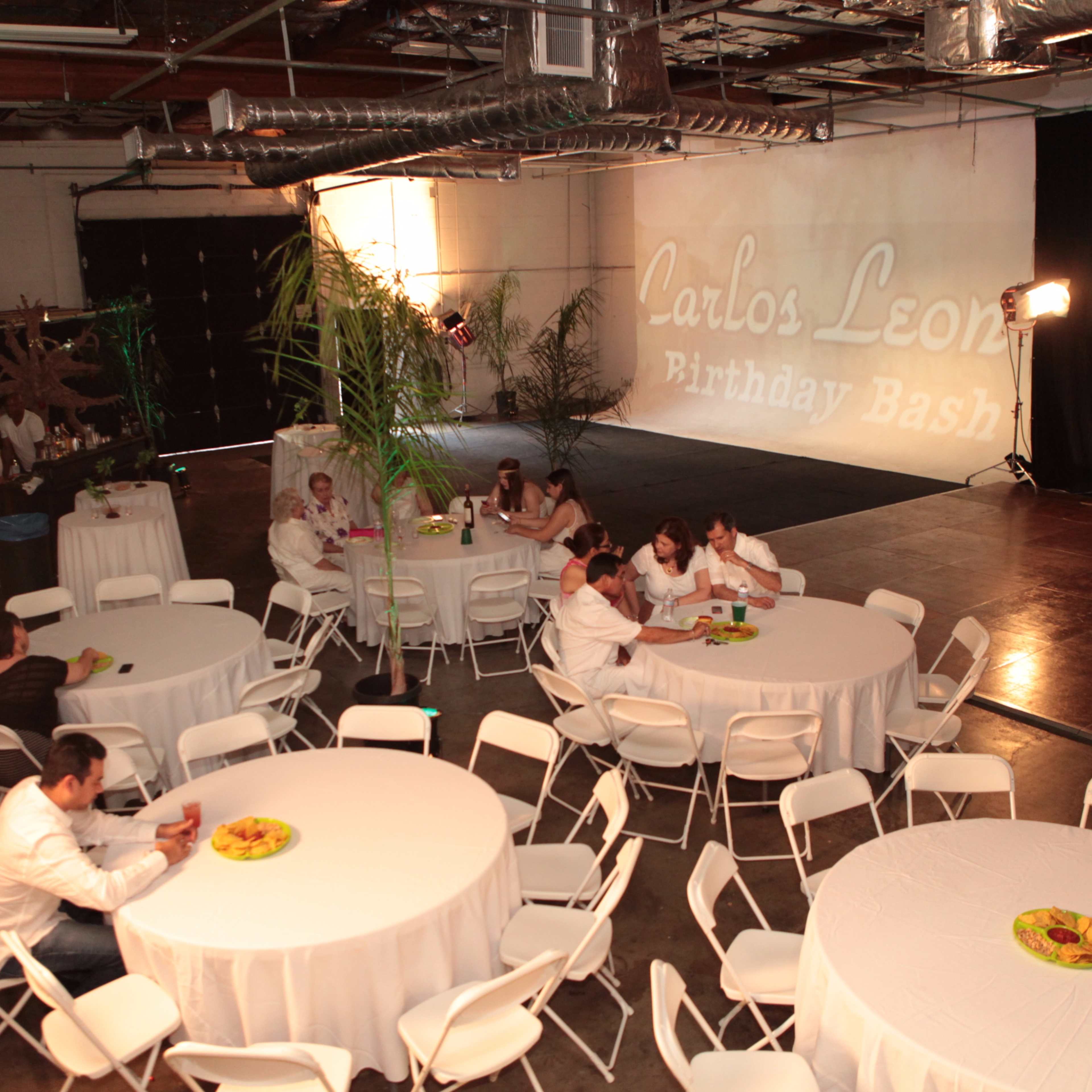 A spacious event venue features white round tables with chairs, a birthday banner reading "Carlos Leon Birthday Bash" projected on the wall, and guests seated around tables.
