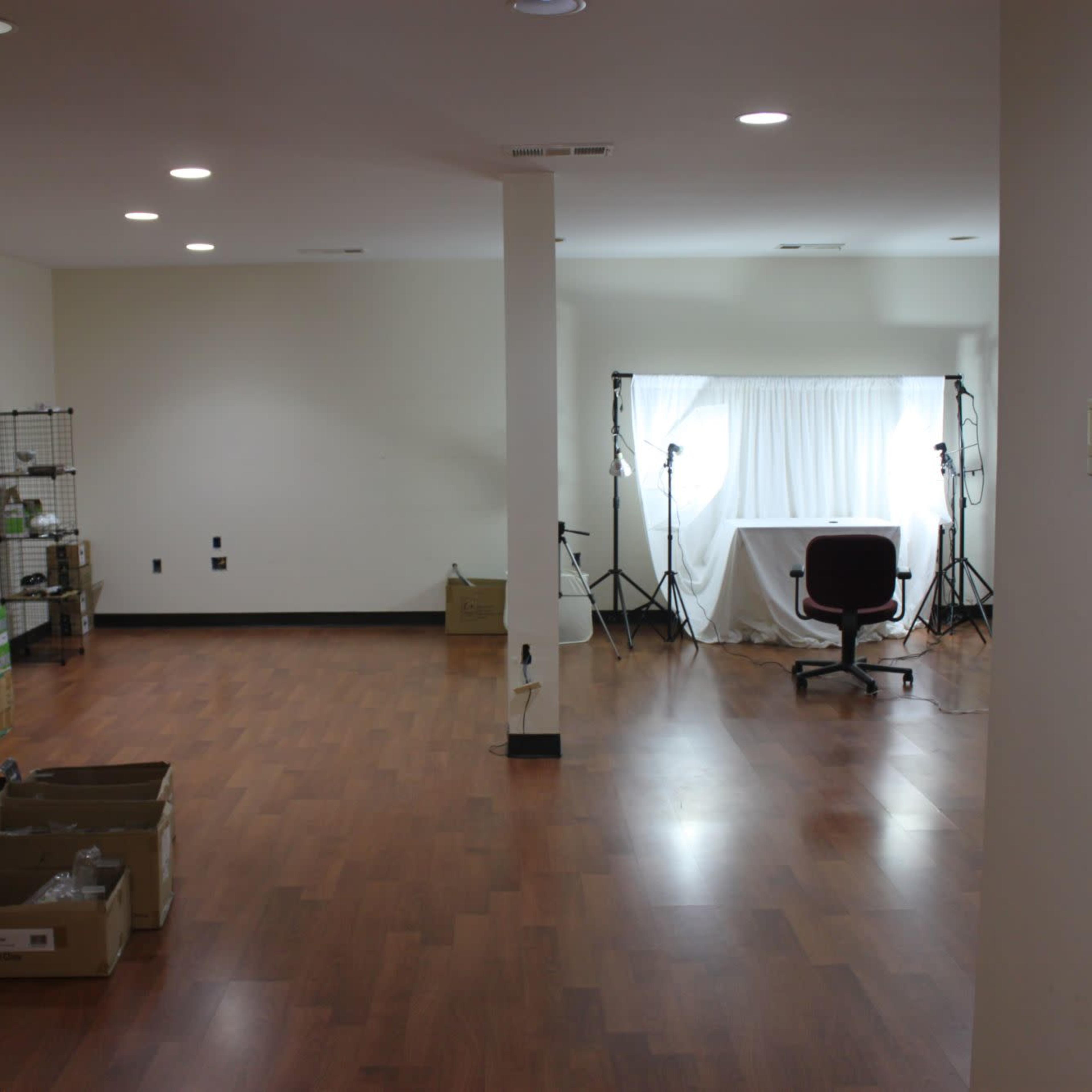 A spacious, empty room with wood flooring, a window covered by light curtains, and a chair positioned near photography equipment on stands.