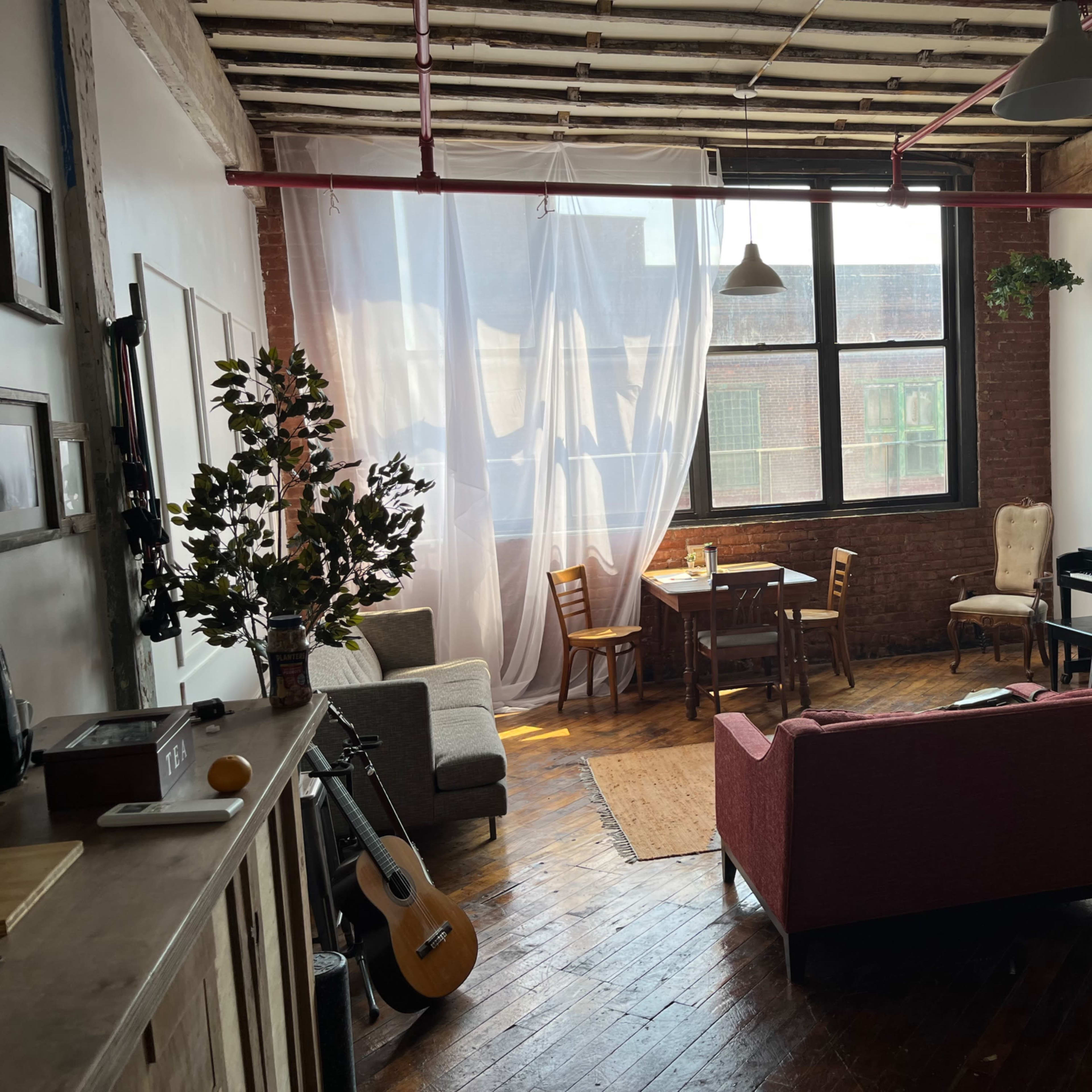 A spacious, bright living room with wooden floors, featuring a piano, a dining table, and various seating arrangements near large windows.