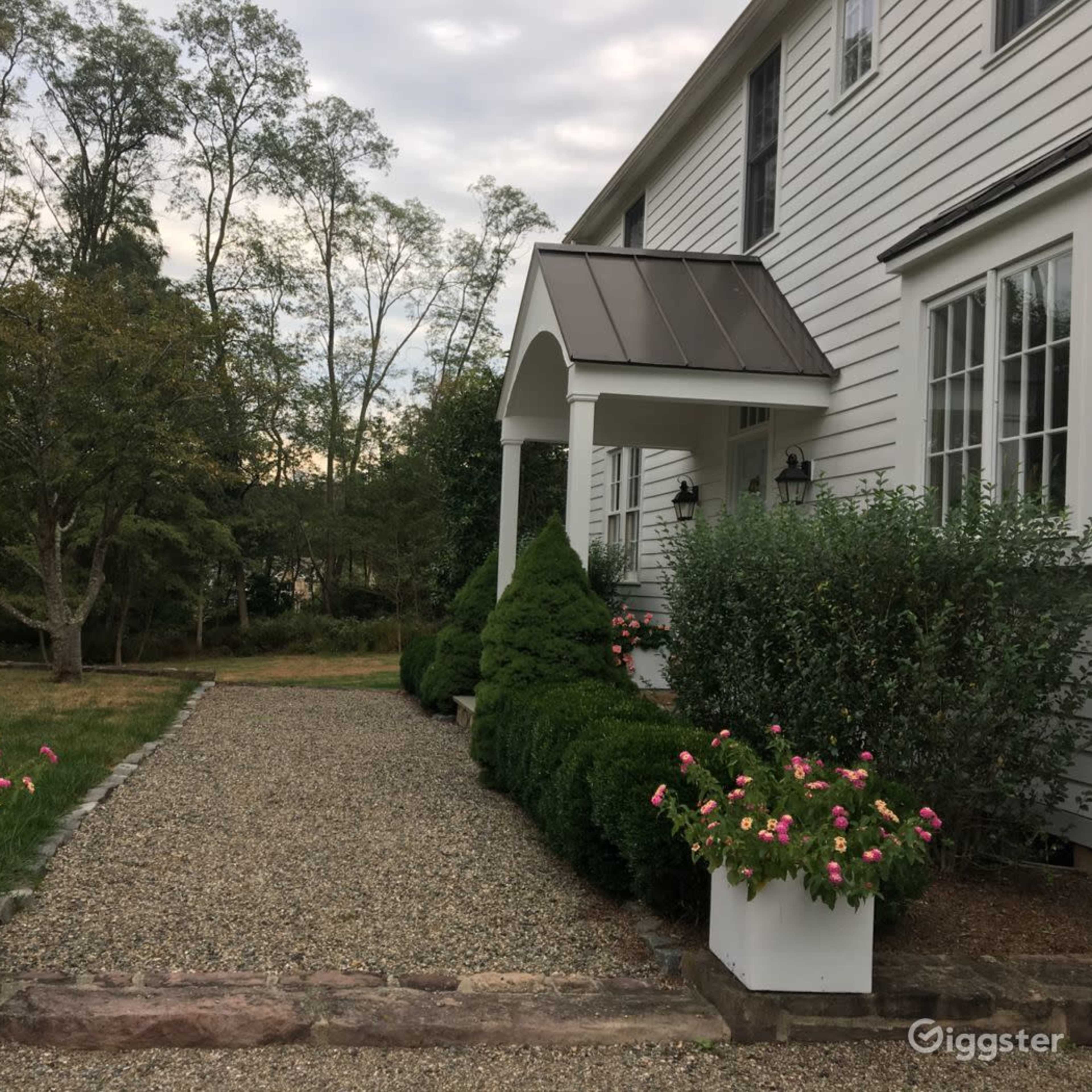 A gravel pathway lined with planters of pink flowers leads to the entrance of a white house with a covered porch.