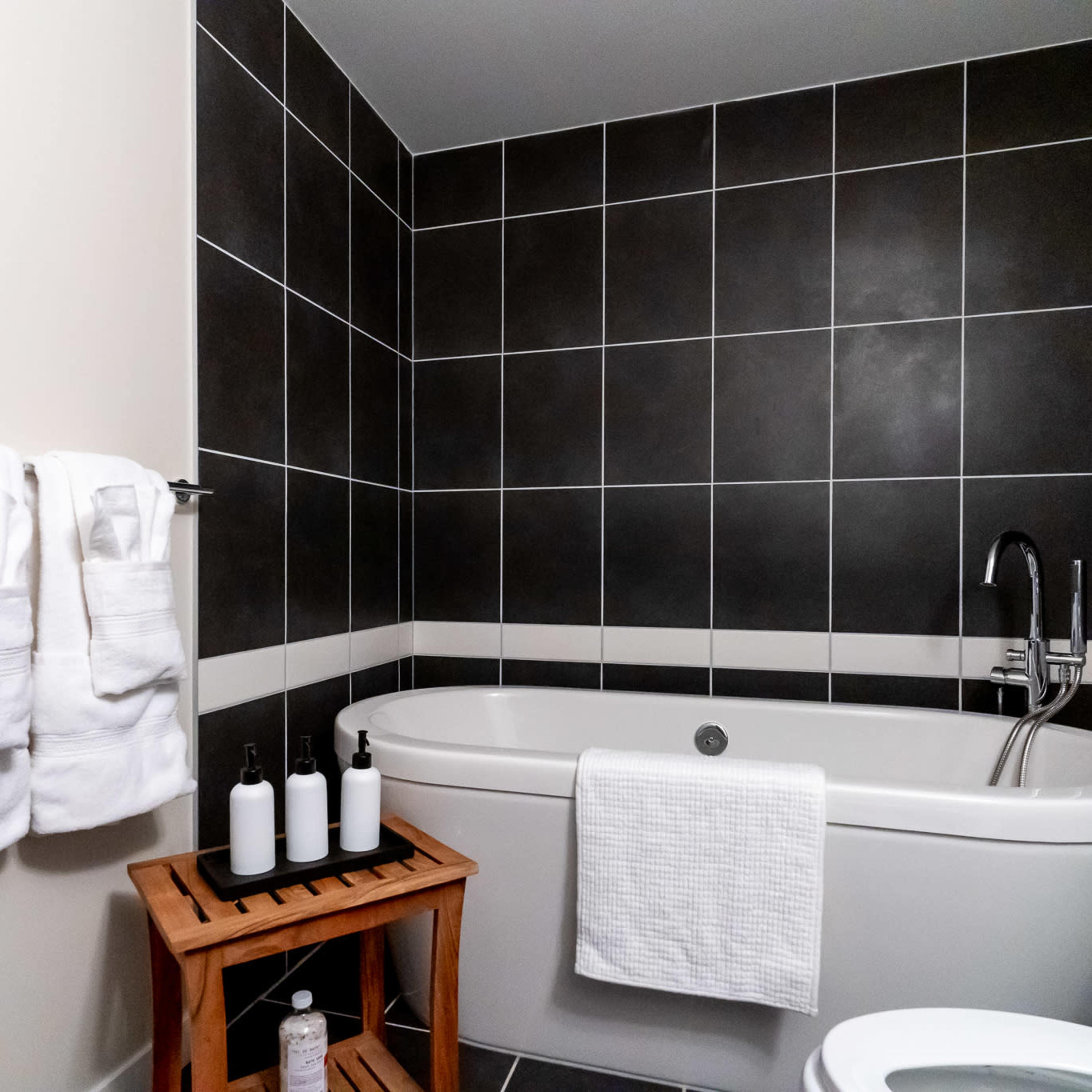 A modern bathroom features a white bathtub, a wooden stool with toiletries, and neatly hung towels on a towel rack.
