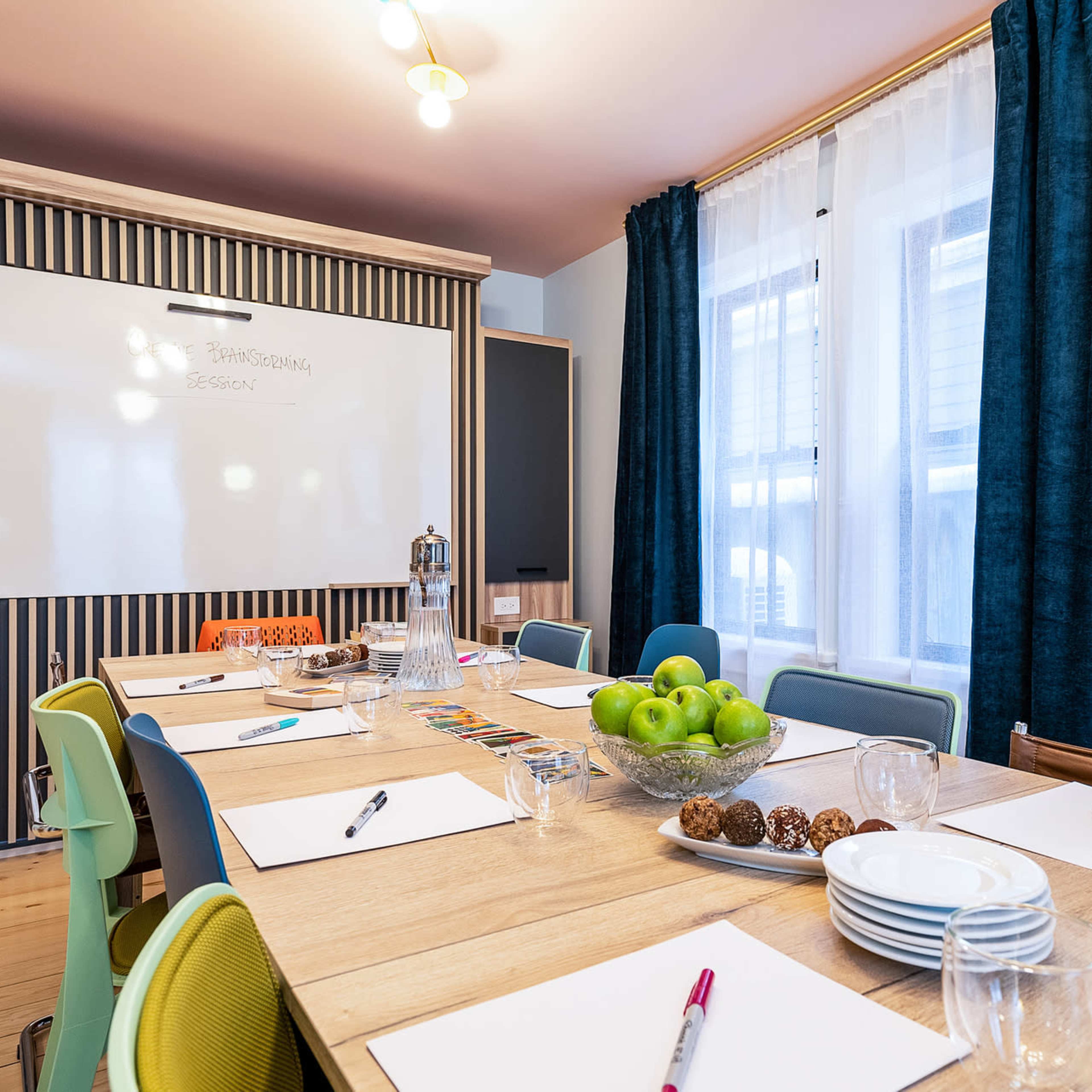 A conference room is set up with a long wooden table, colorful chairs, and a whiteboard in the background, along with supplies for a meeting.