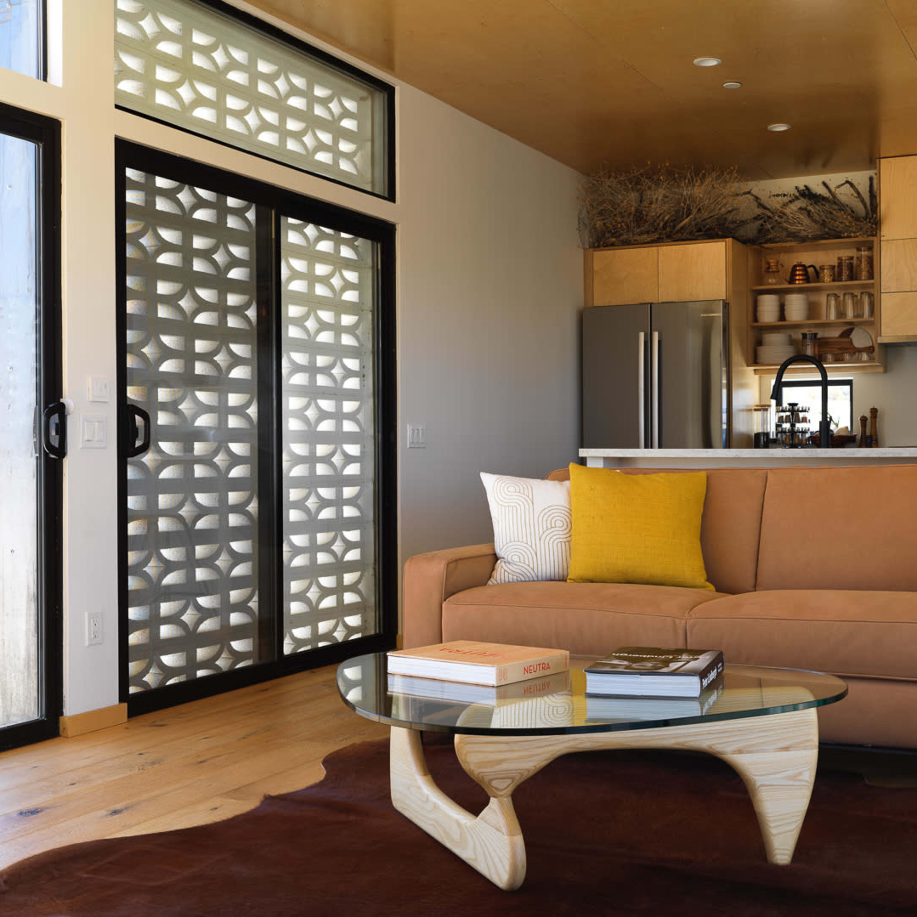 A modern living room featuring a brown sofa, a glass coffee table on a dark rug, and a kitchen area with wooden cabinets in the background.