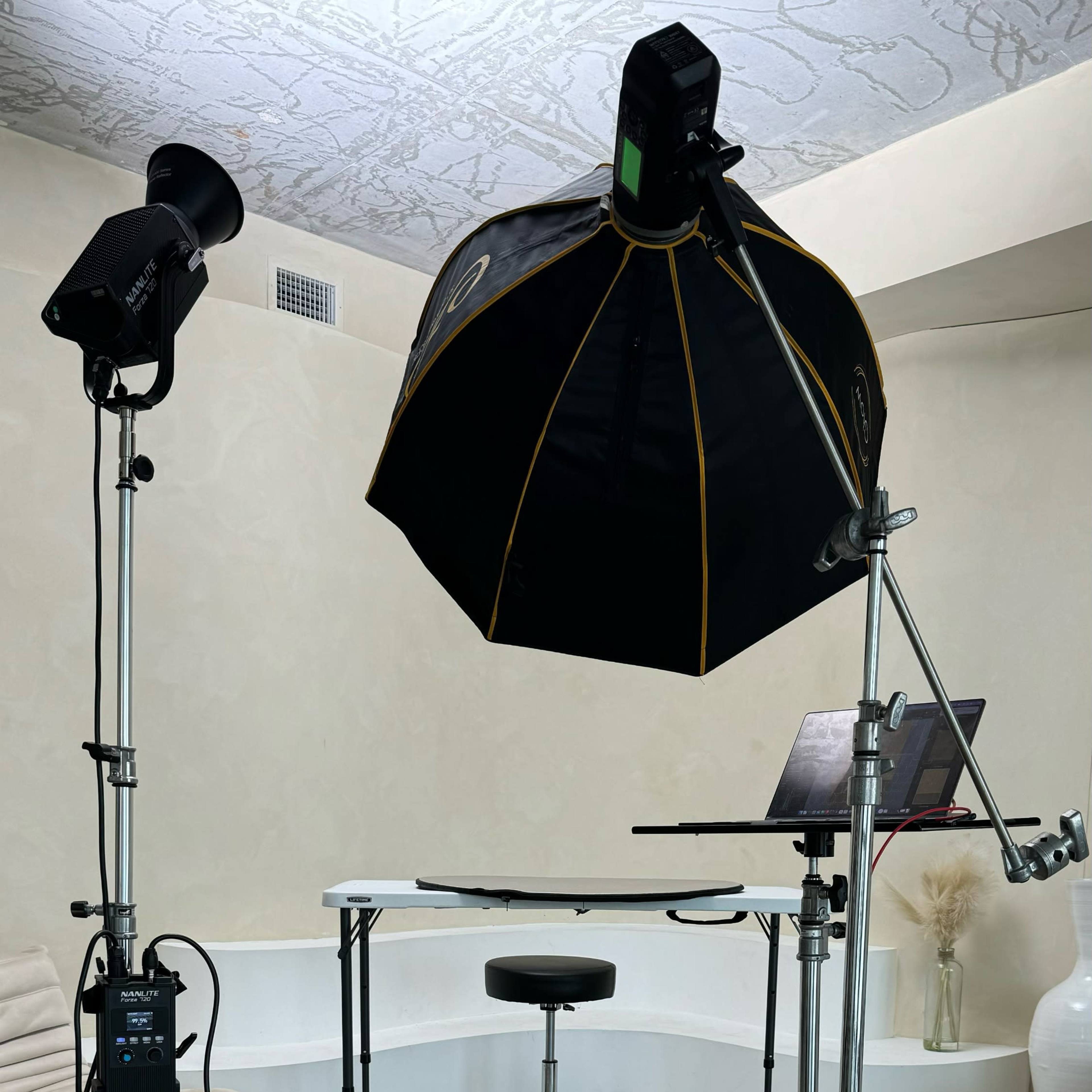 A photography setup with a camera on a tripod, a light umbrella softbox, a stool, and various equipment arranged on a studio floor.