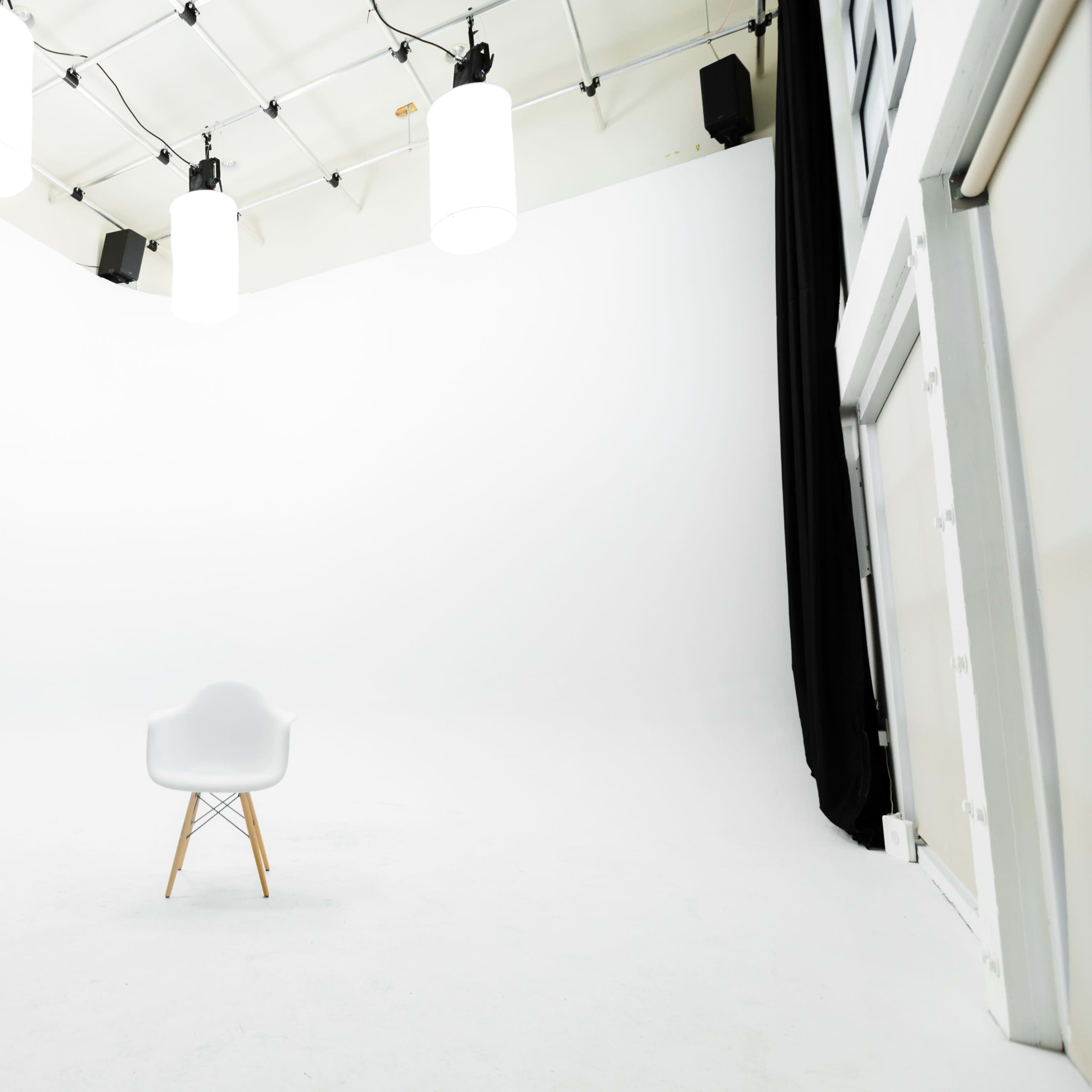 An empty studio space with a single white chair centered on a white floor, illuminated by several hanging lights.