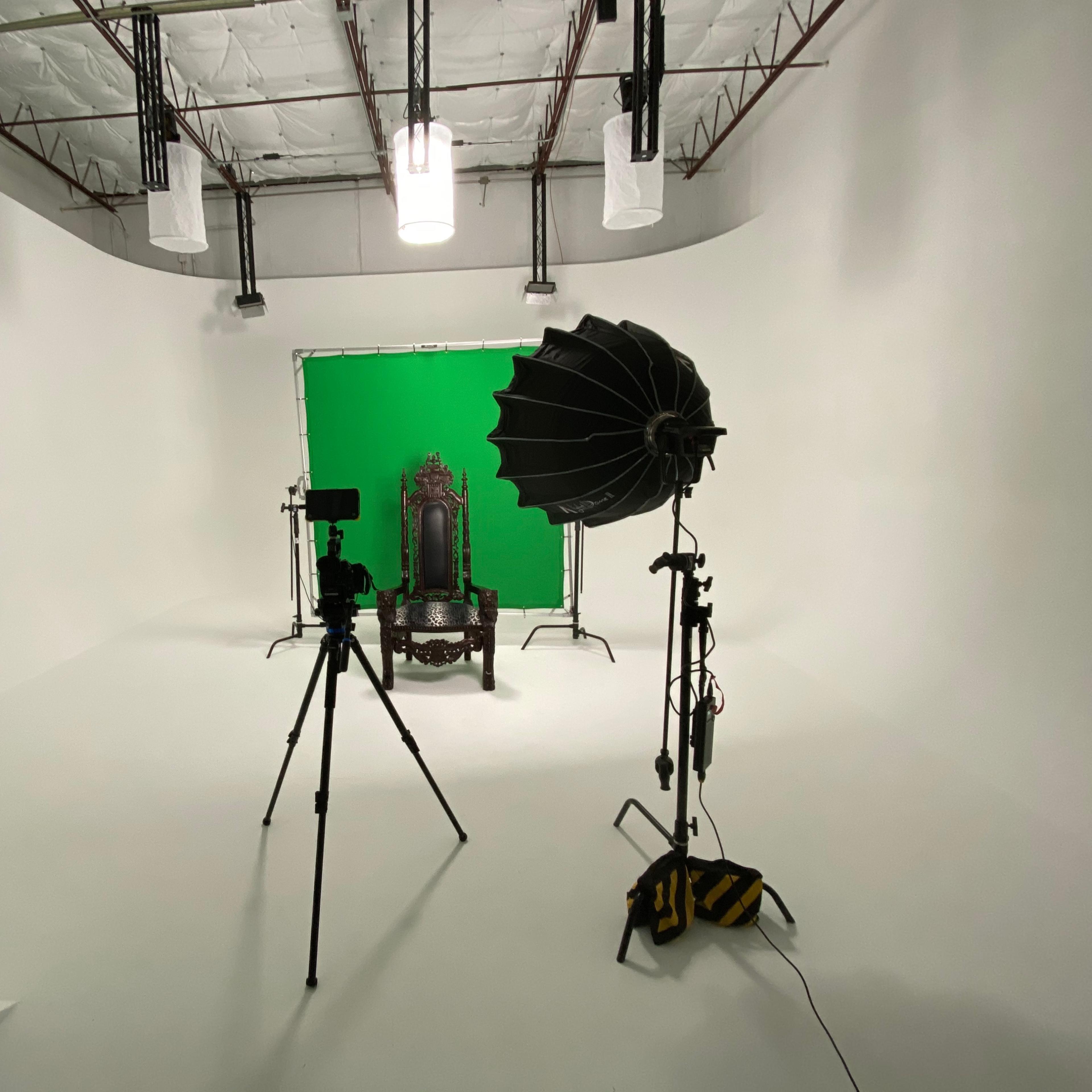 A photography studio with a white backdrop, a green screen set up, a vintage chair, and lighting equipment arranged around a camera on a tripod.
