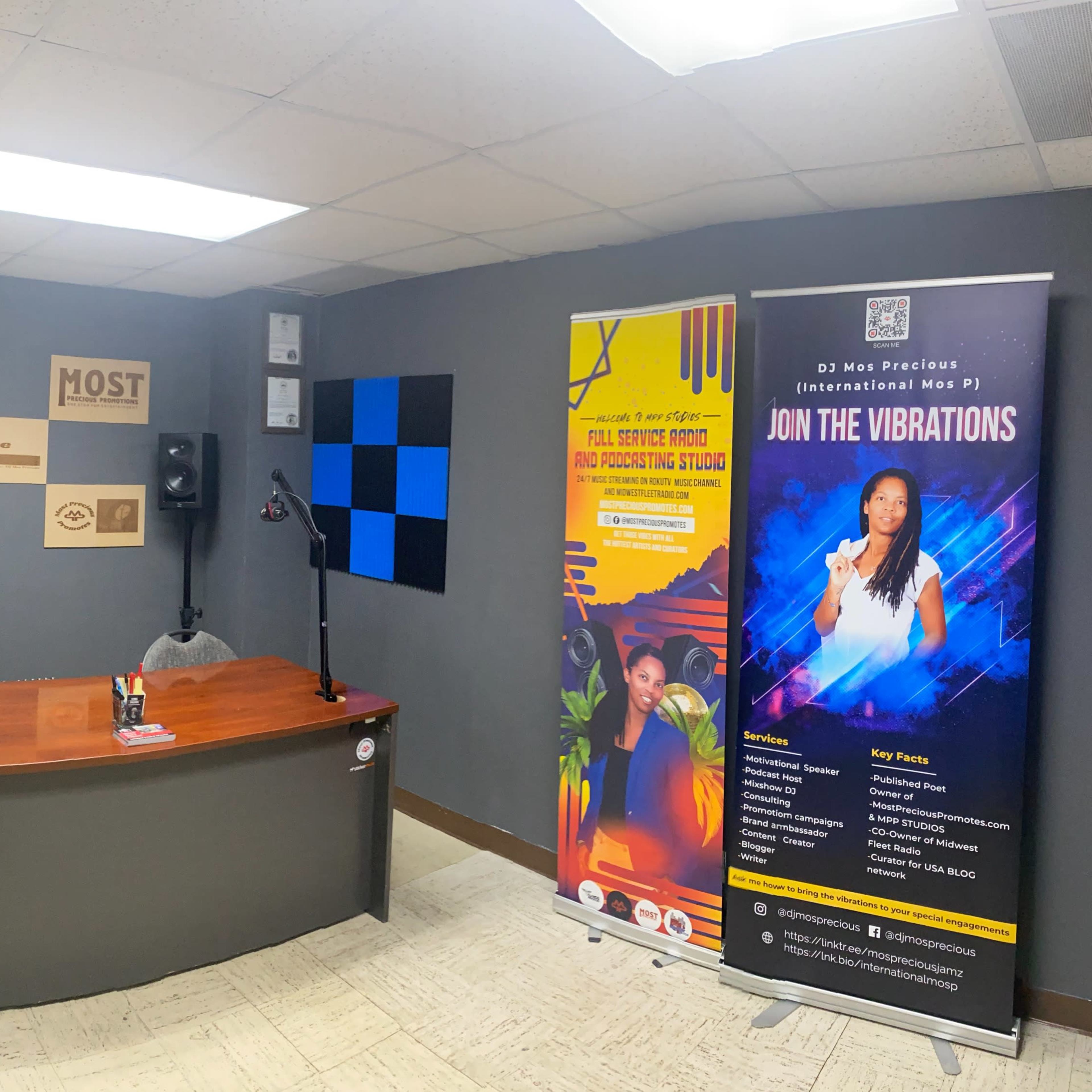 A radio studio featuring a wooden desk with microphones, soundproofing panels on the walls, and promotional banners on either side.