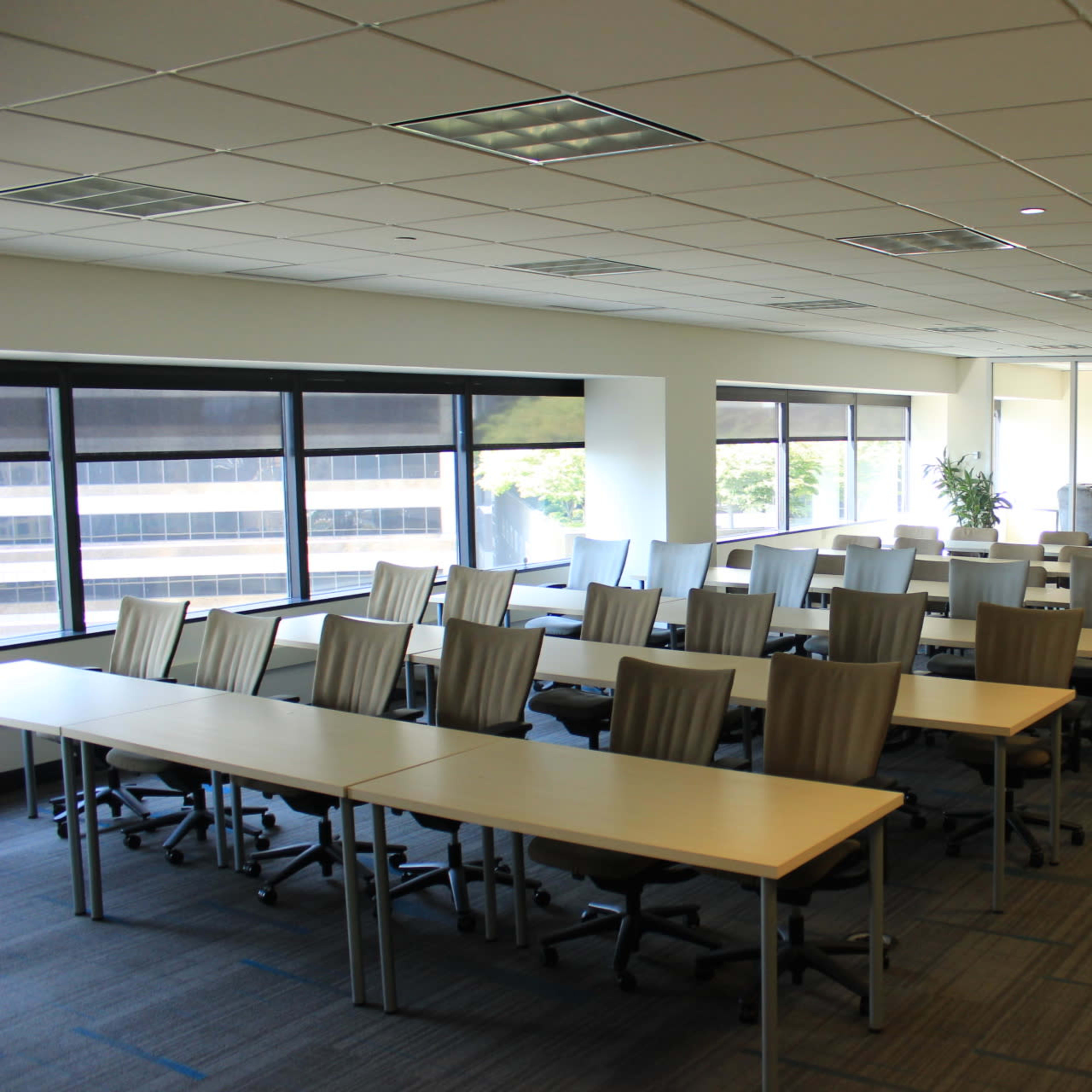 A conference room with rows of tables and chairs, large windows offering views of a cityscape, and a whiteboard at one end.