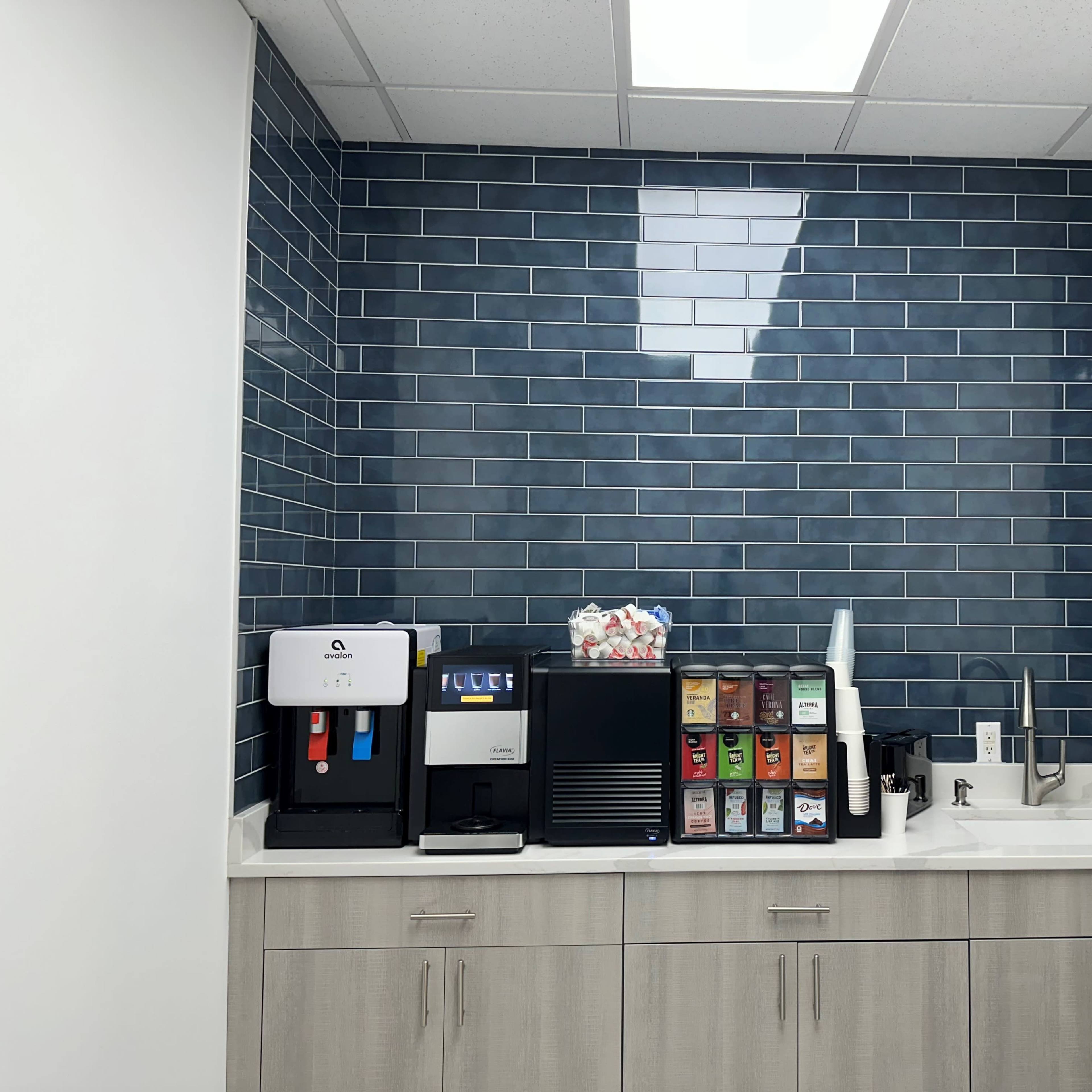 A kitchenette area featuring a countertop with a coffee machine, a beverage dispenser, and a sink against a blue tiled wall.