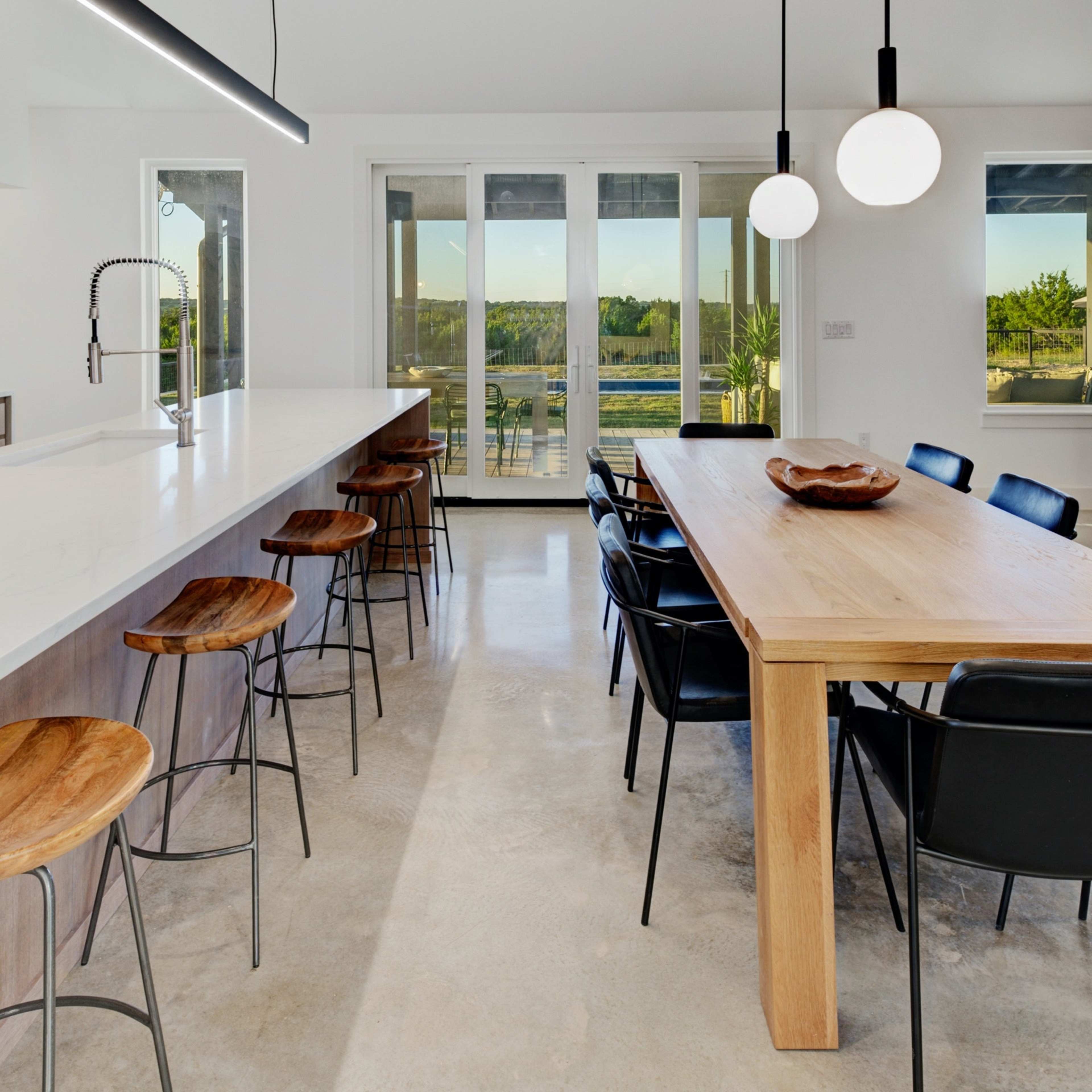 A modern kitchen and dining area with a long wooden table, black chairs, a white countertop, and large windows overlooking a view.