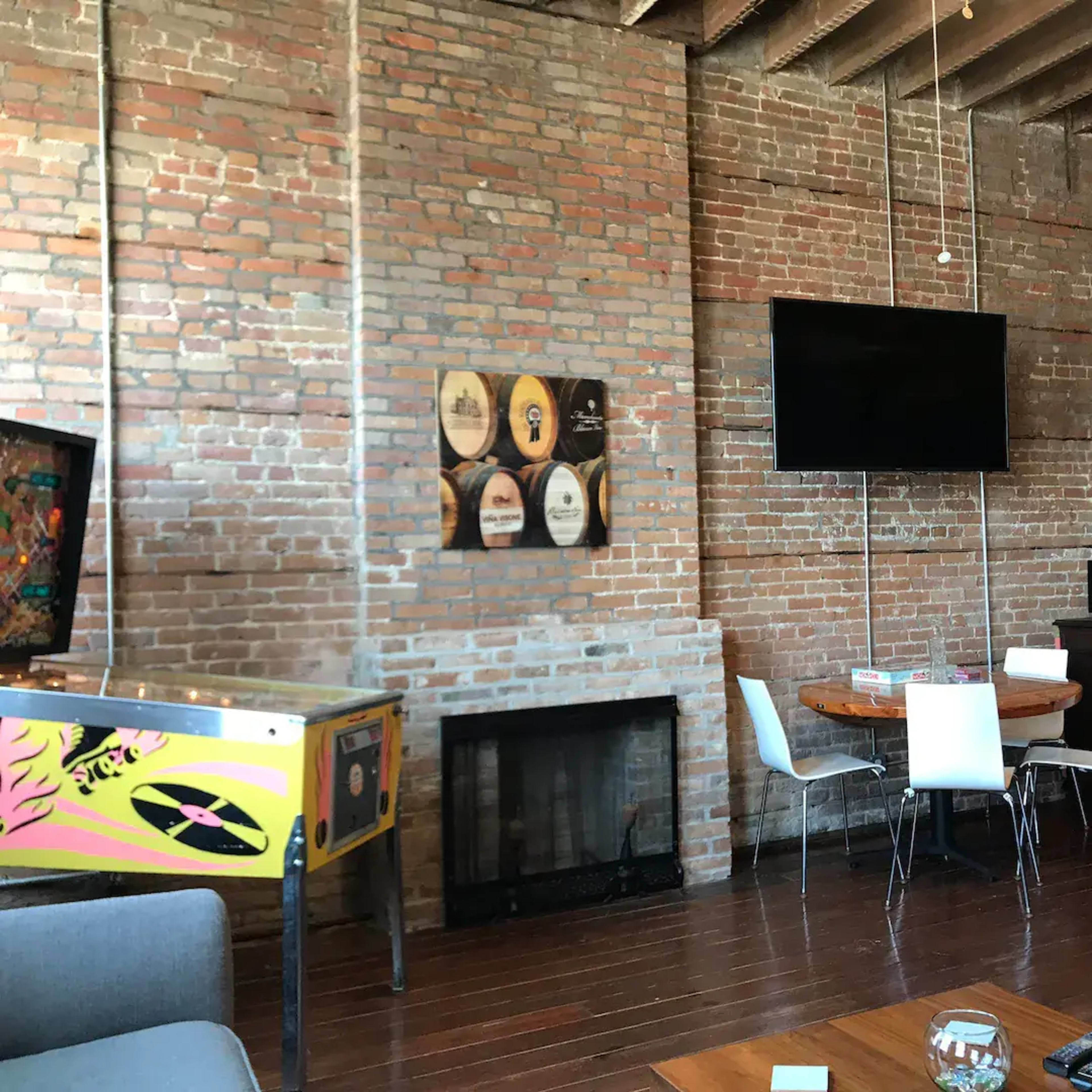 An interior space with exposed brick walls, a wooden floor, a round dining table with white chairs, a wall-mounted television, and a vintage arcade machine.