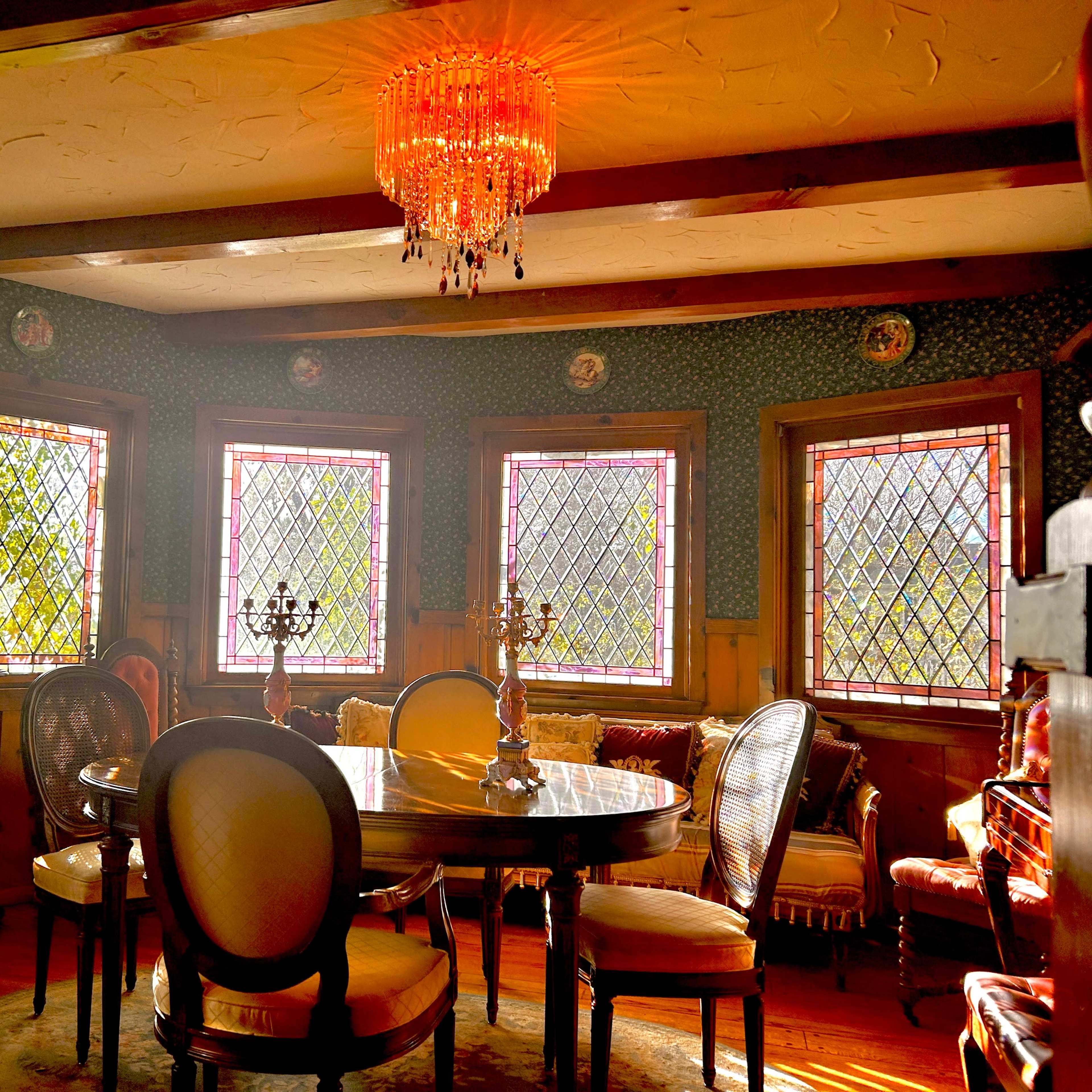 A dining room with a round table surrounded by upholstered chairs, illuminated by a chandelier and featuring large, stained glass windows.