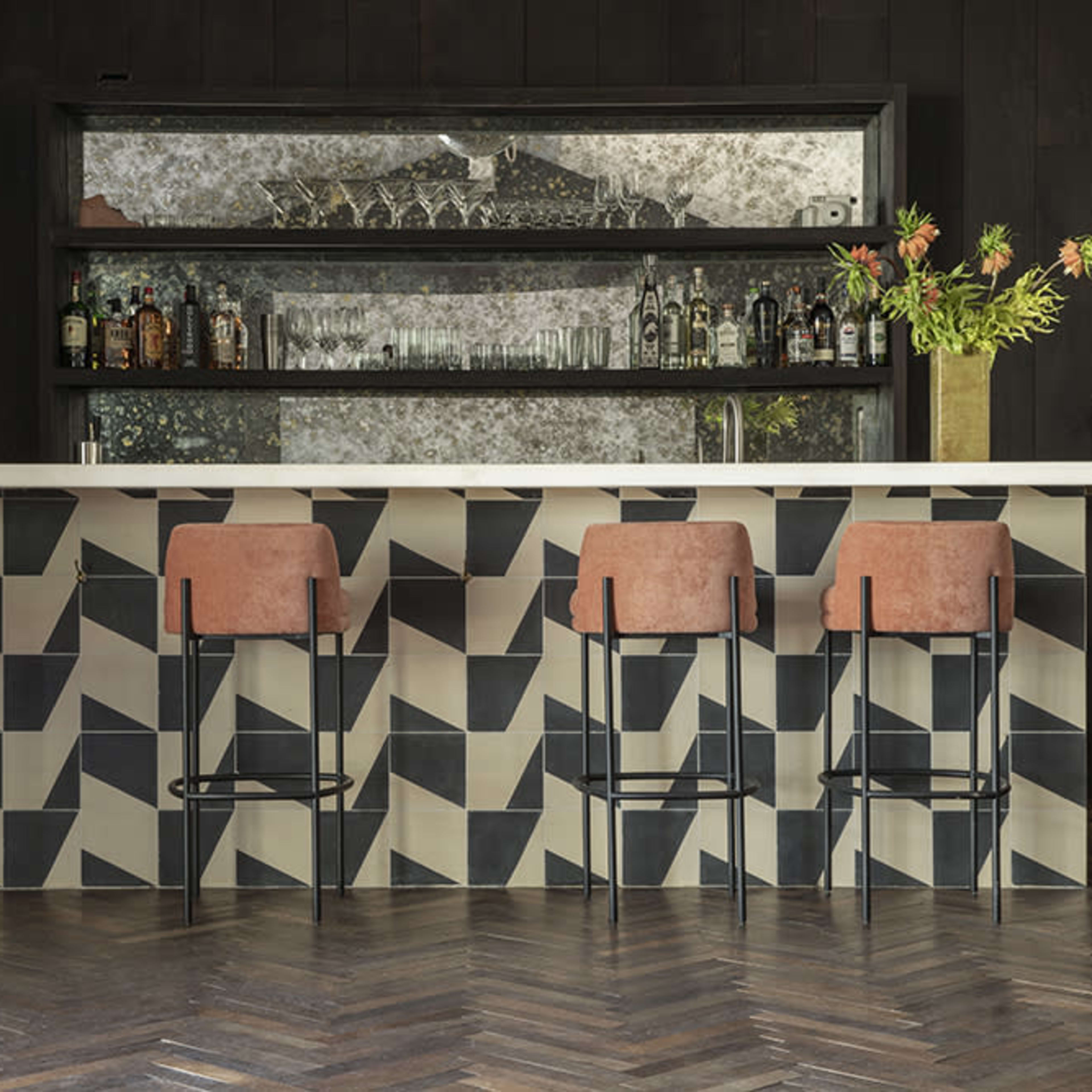 A modern bar with a geometric-patterned countertop, four upholstered stools, and a mirrored back display containing various bottles.