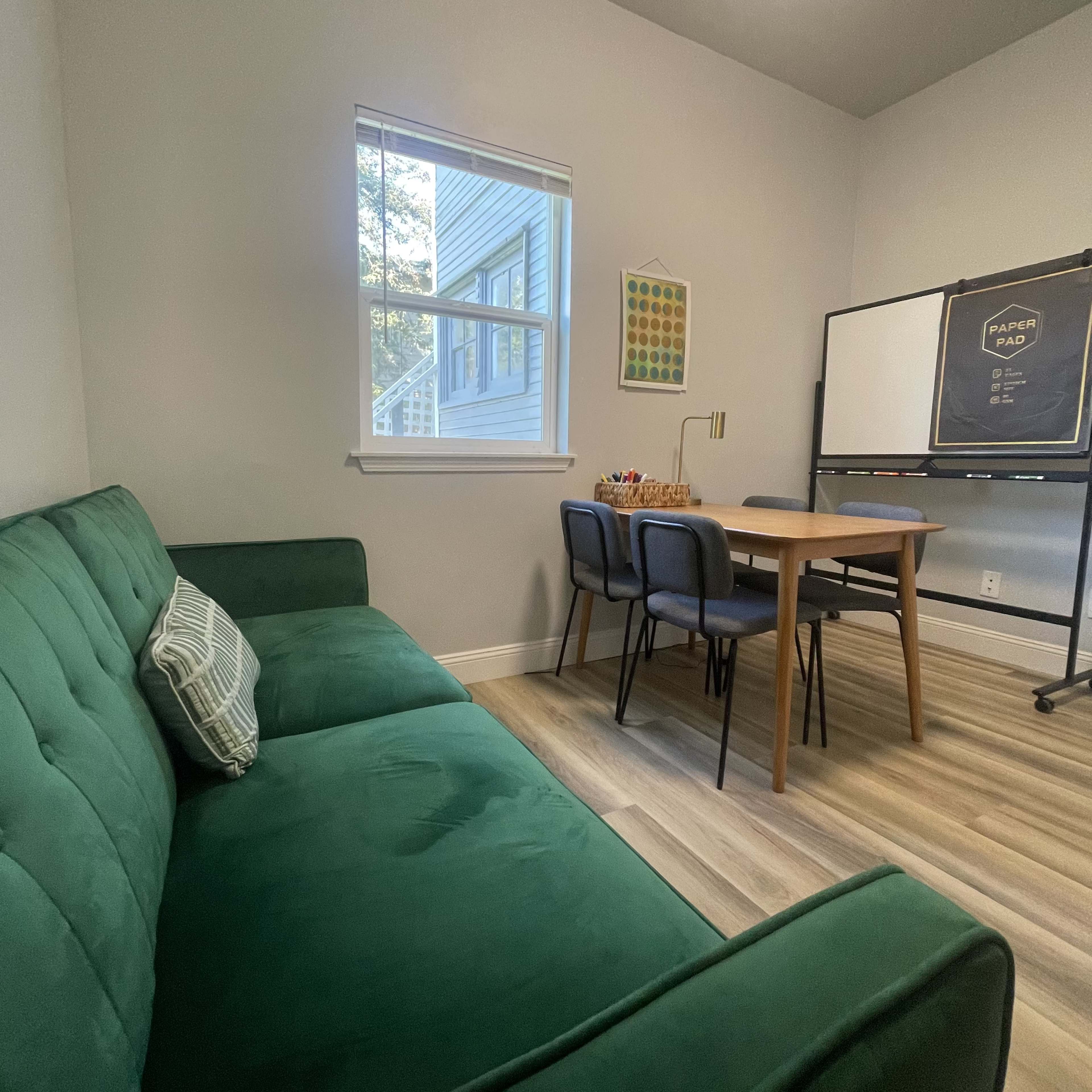 A small room features a green sofa, a wooden table with chairs, and a window with natural light.