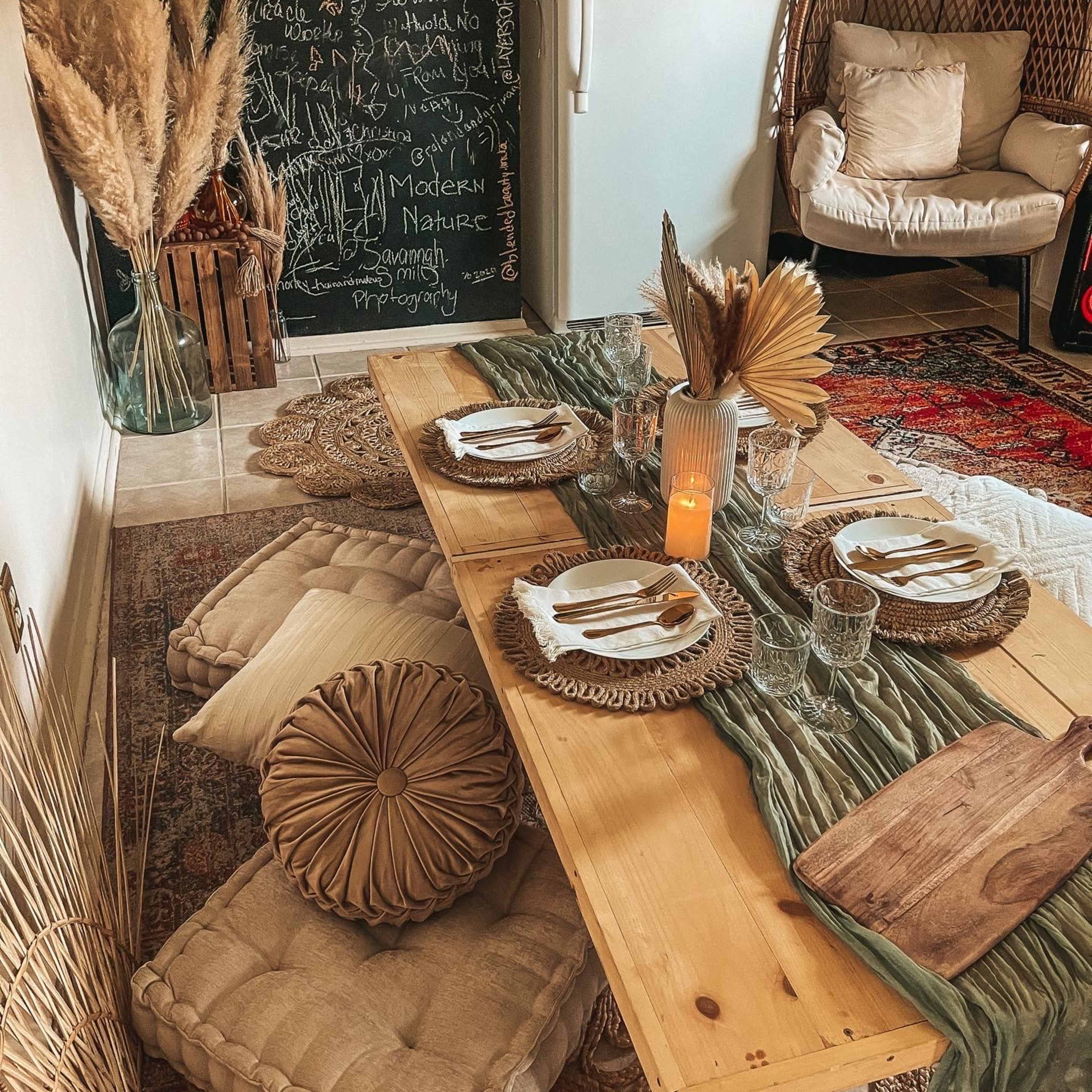 A dining space features a wooden table set with plates and glassware, surrounded by cushions, a chalkboard wall, and decorative elements like pampas grass and a glowing candle.