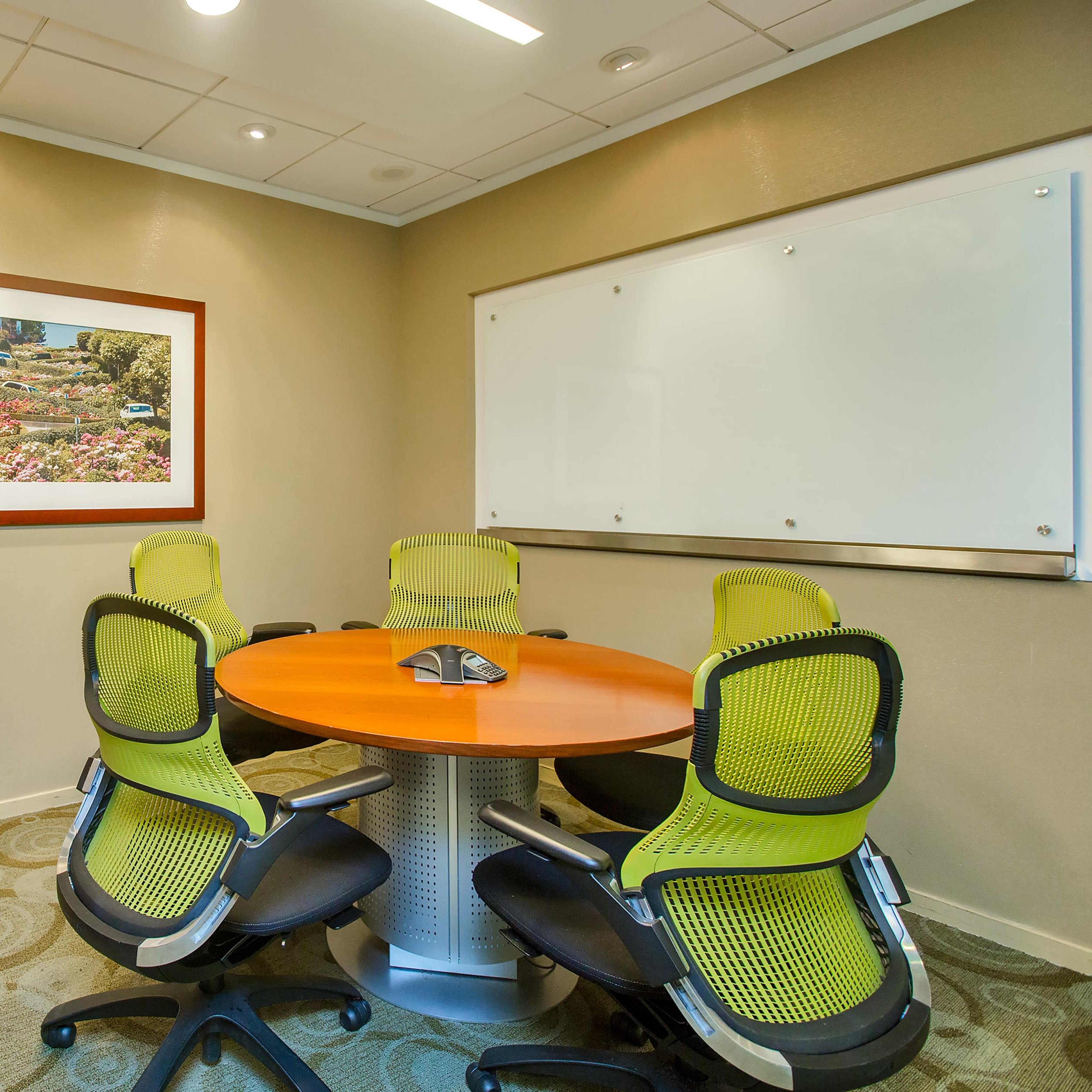 A small conference room with a circular orange table surrounded by four green ergonomic chairs, a whiteboard, and a framed picture on the wall.