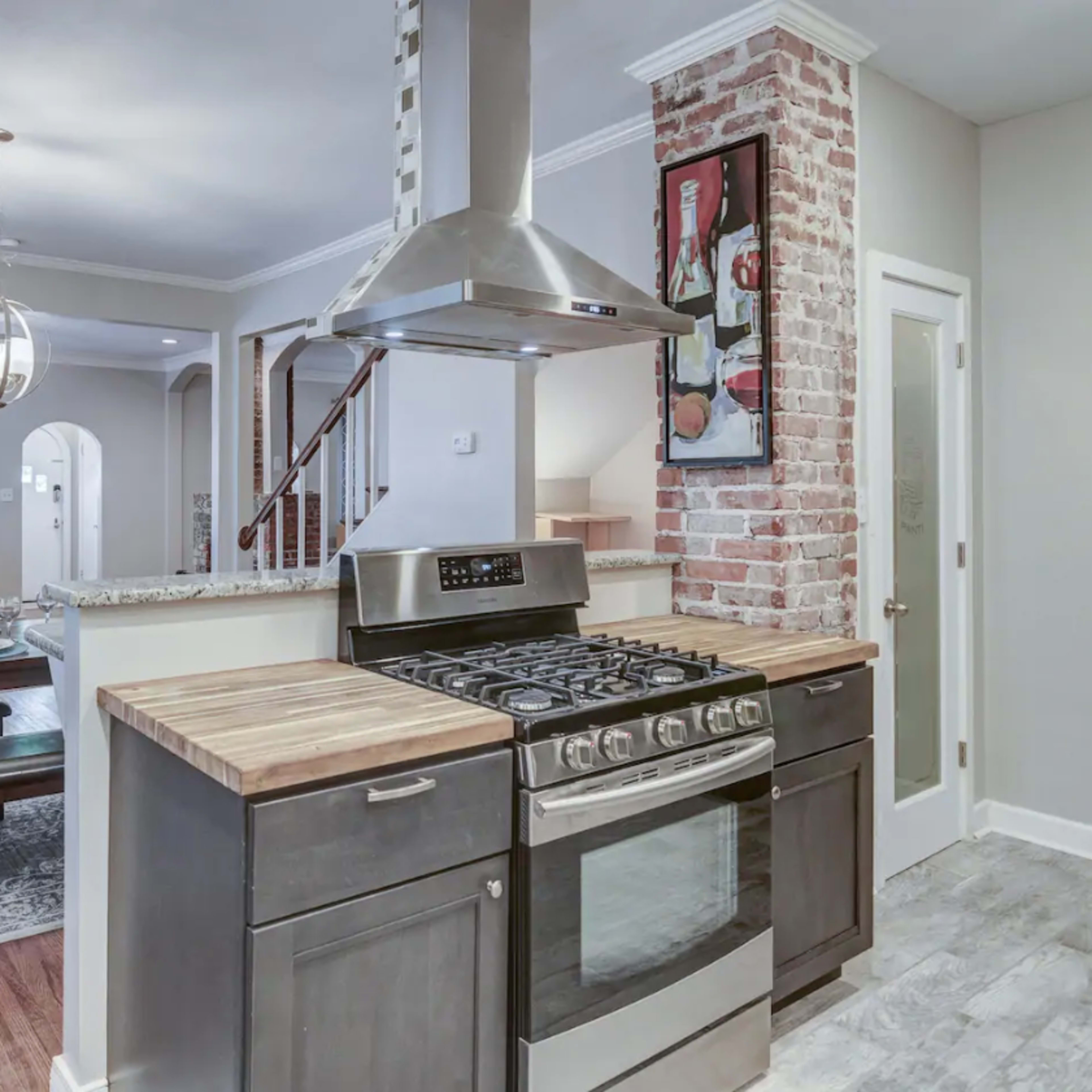 A modern kitchen with a gas stove, wooden countertops, and an exposed brick wall, adjacent to a dining area with a table set for four.