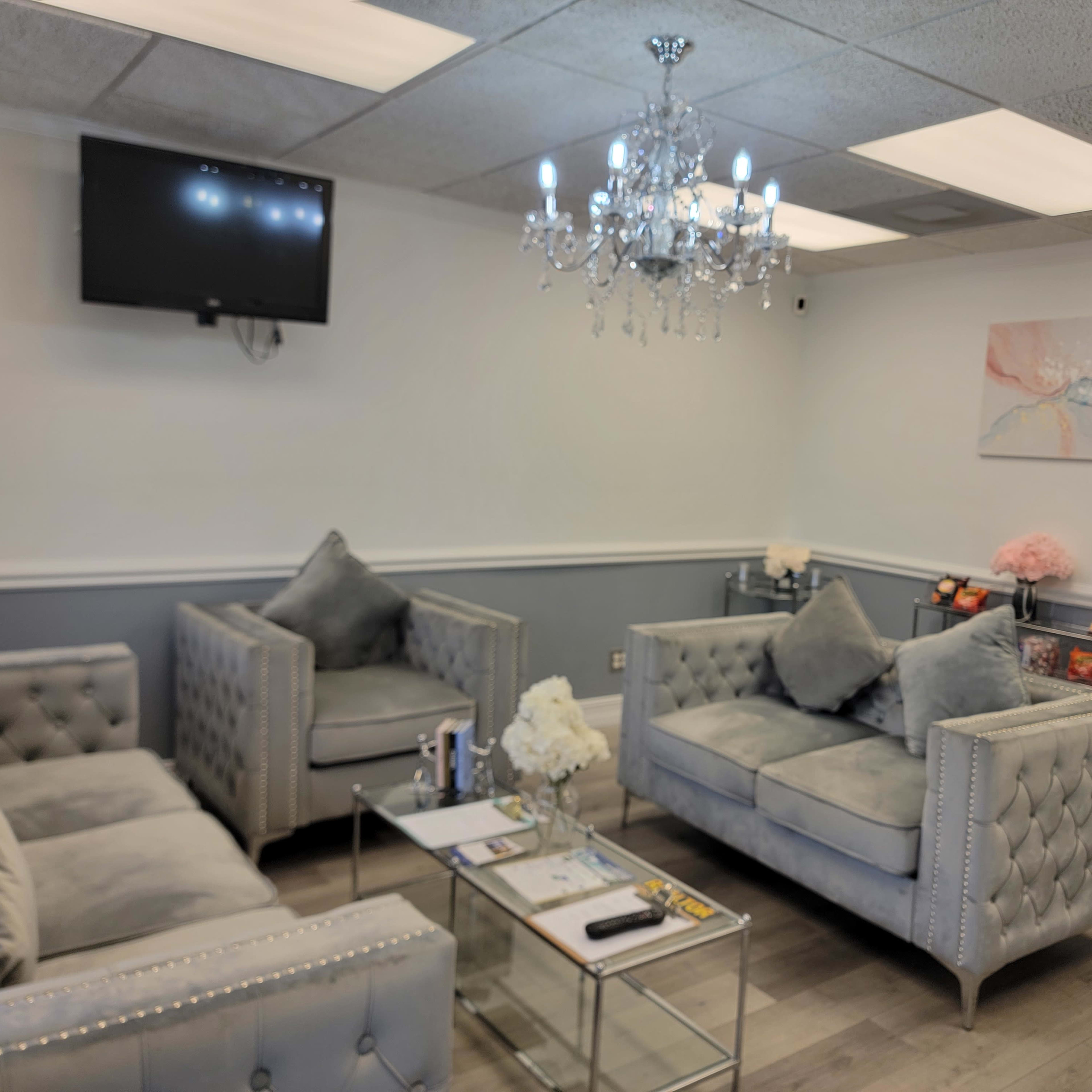 A well-furnished waiting area featuring plush gray sofas, a glass coffee table, and a chandelier, with a television mounted on the wall.