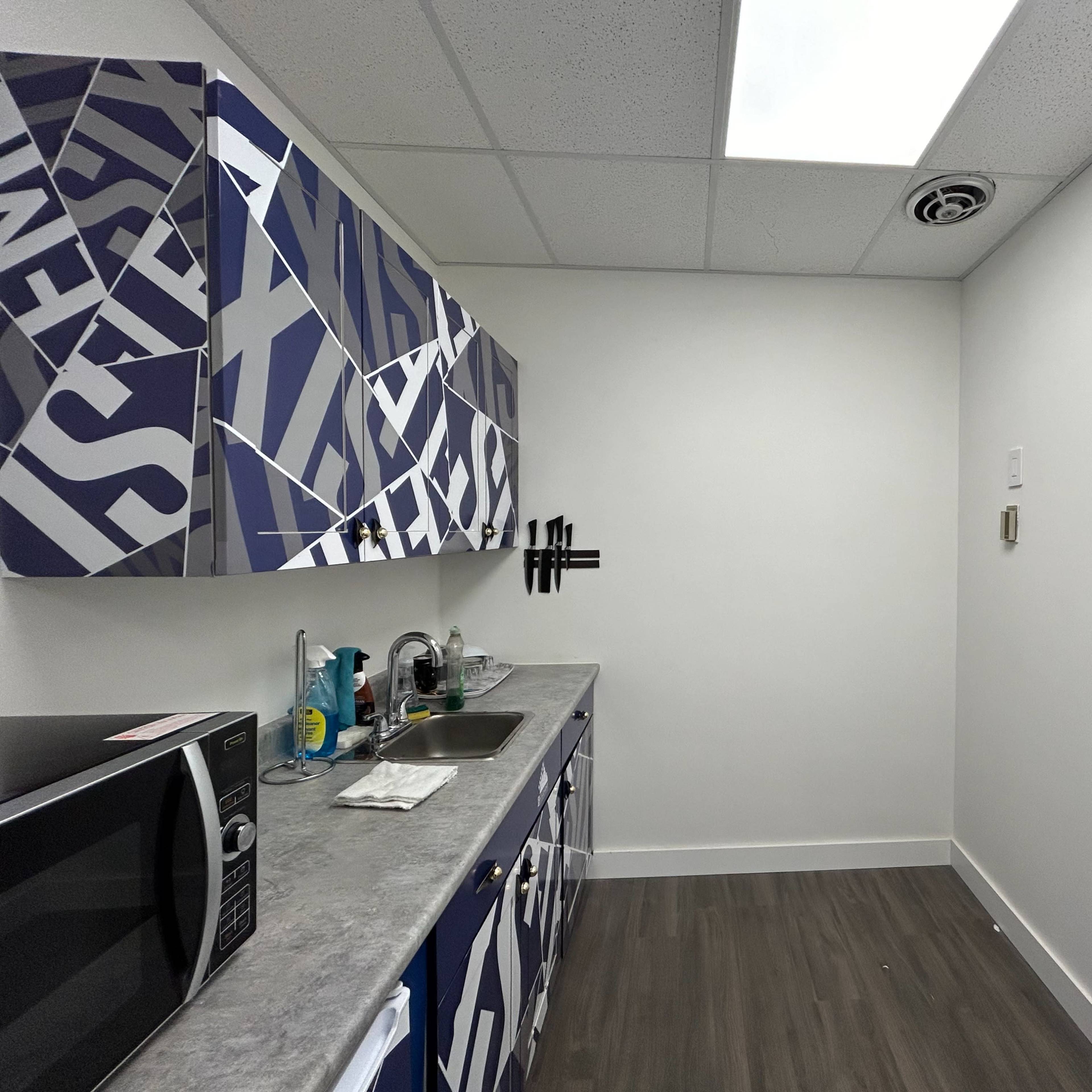 A small kitchen area with blue cabinets, a sink, a microwave, and a wall-mounted device on the right.