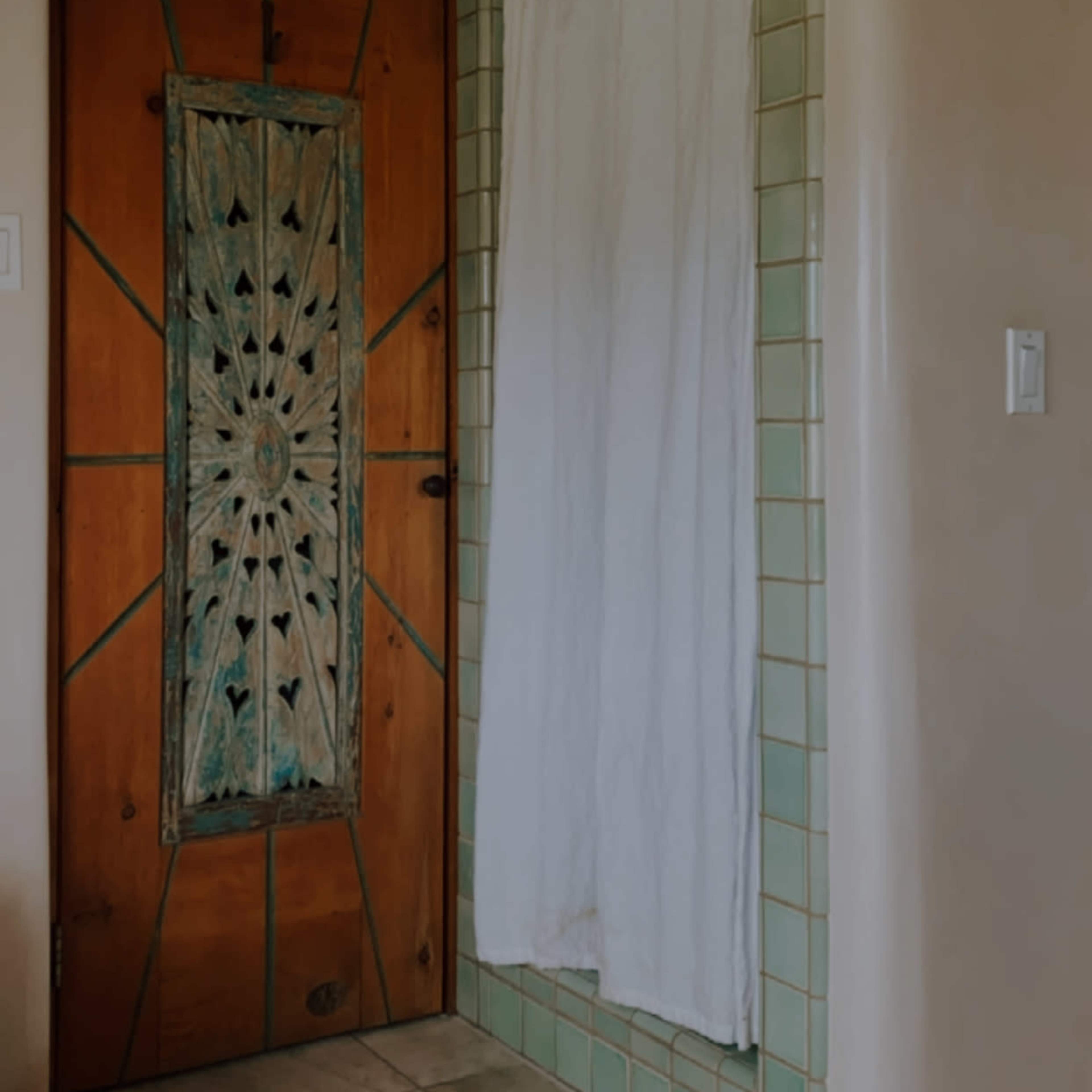 A wooden door with intricate carvings stands adjacent to a tiled shower area covered by a white curtain.