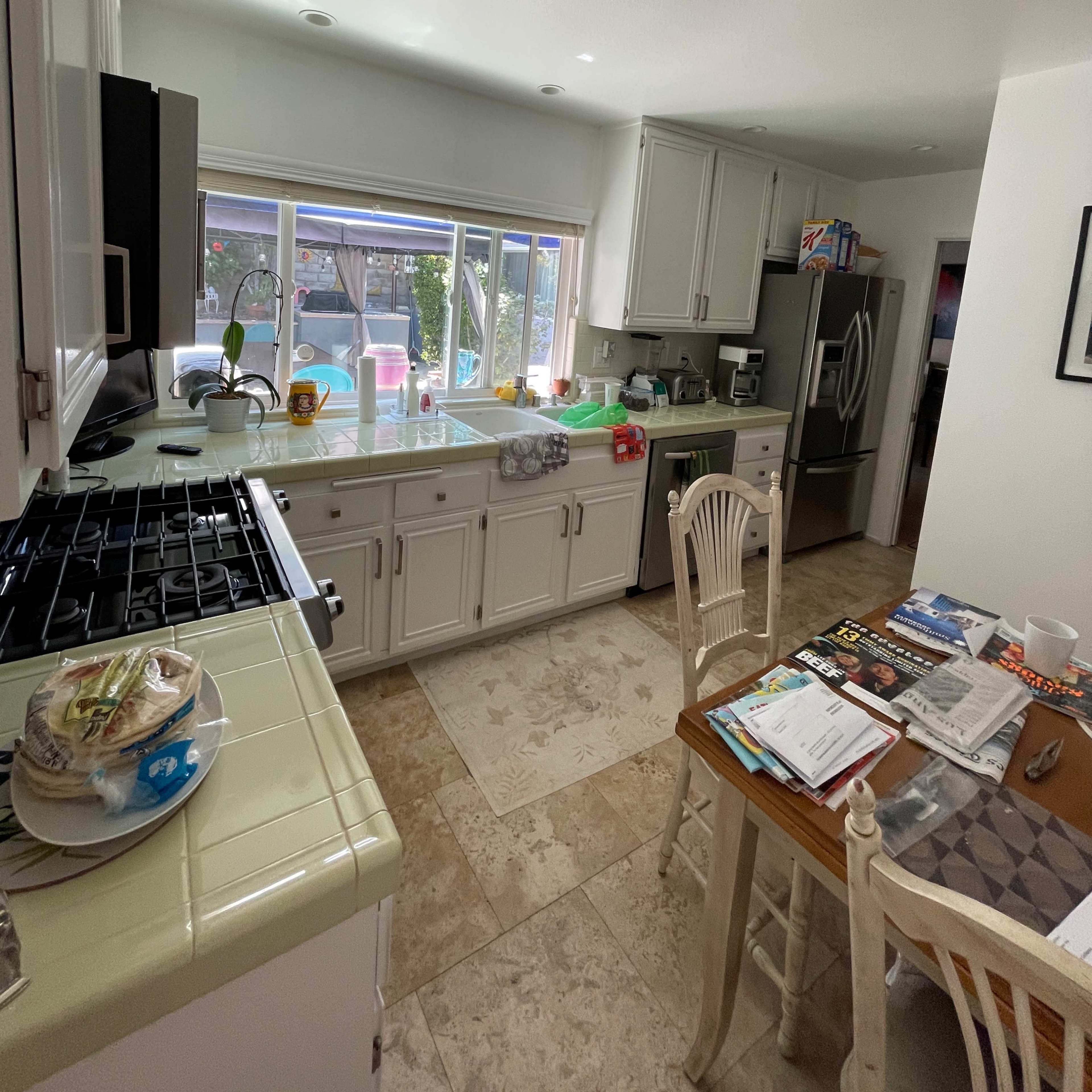 A kitchen with white cabinets and a tiled floor, featuring a stove, refrigerator, and a dining table cluttered with papers and dishes.