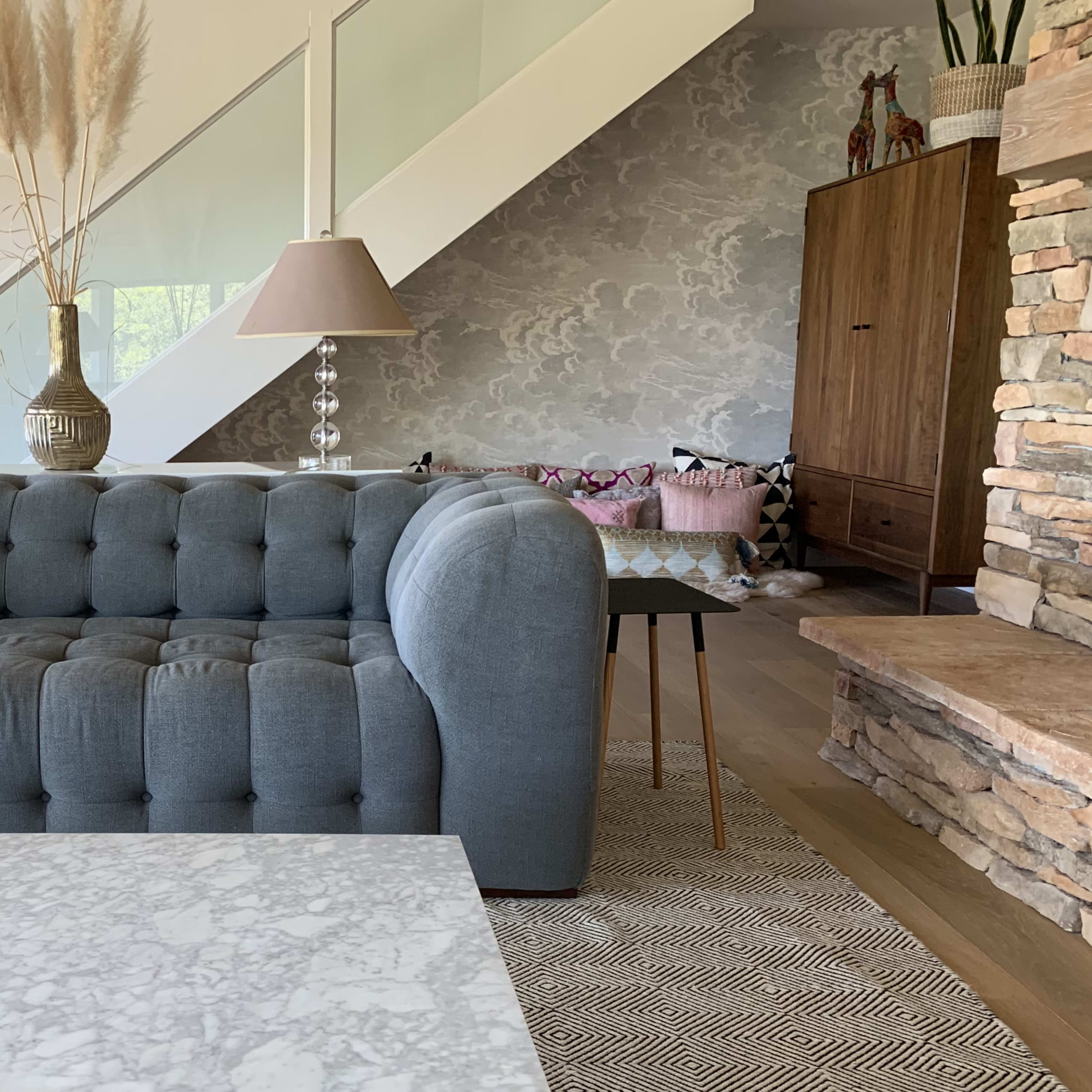 A modern living room featuring a gray tufted sofa, marble coffee table, and a staircase in the background, along with decorative lamps and a wooden cabinet.