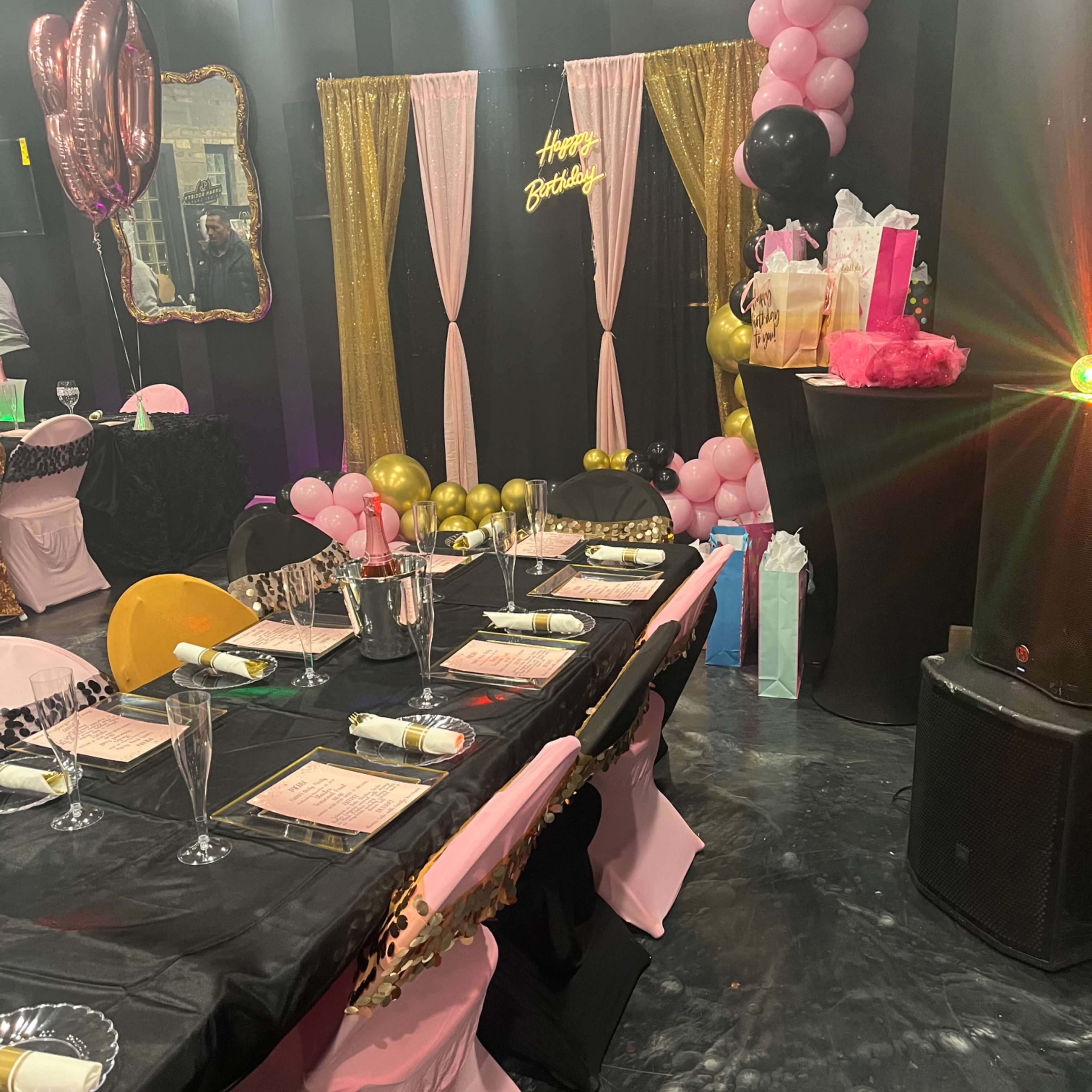 A birthday celebration setup features a decorated table with black tablecloths, pink chairs, and a backdrop of balloons and a "Happy Birthday" sign.