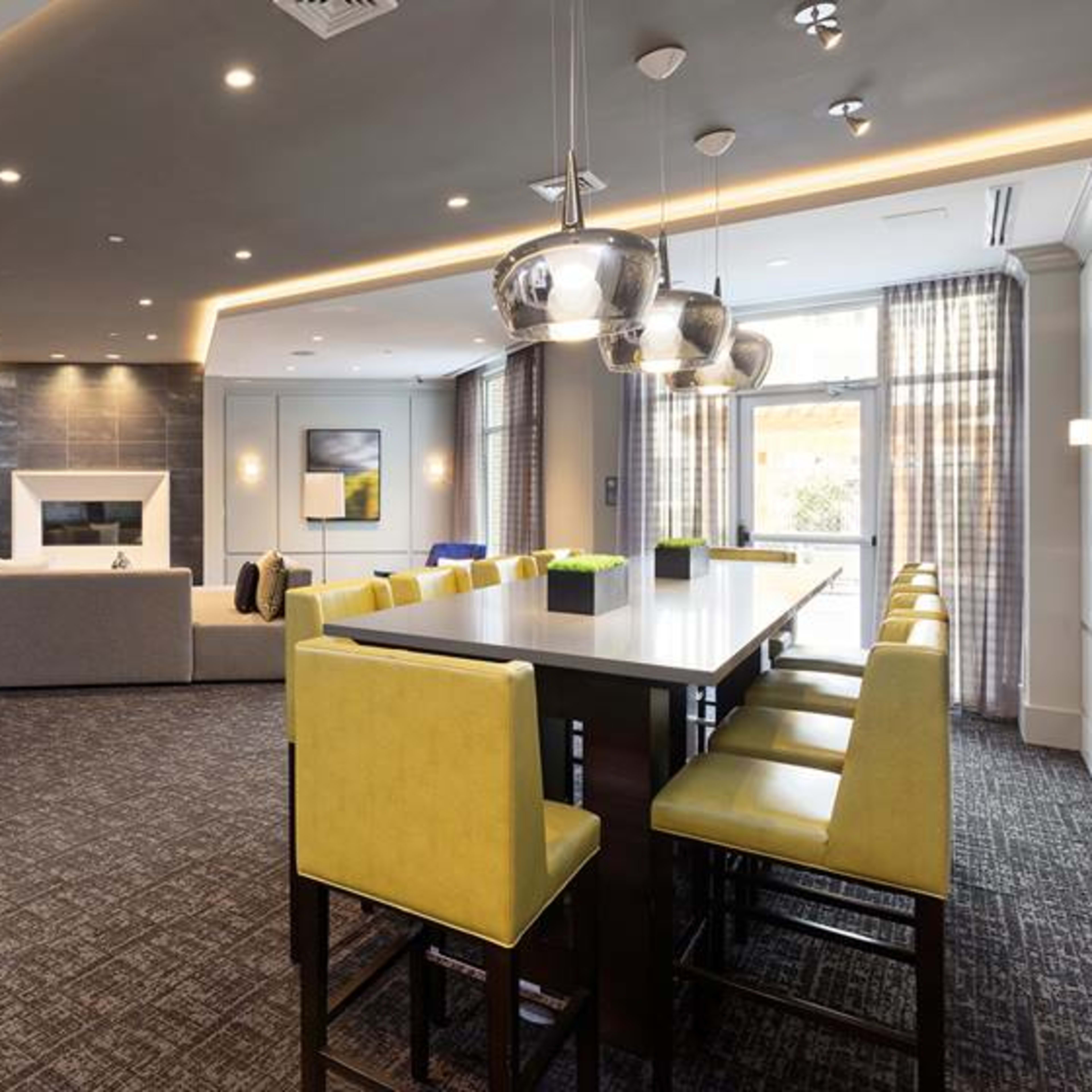 A modern lounge area featuring a large seating arrangement, a dining table with yellow high-back chairs, a fireplace, and a television mounted on the wall.