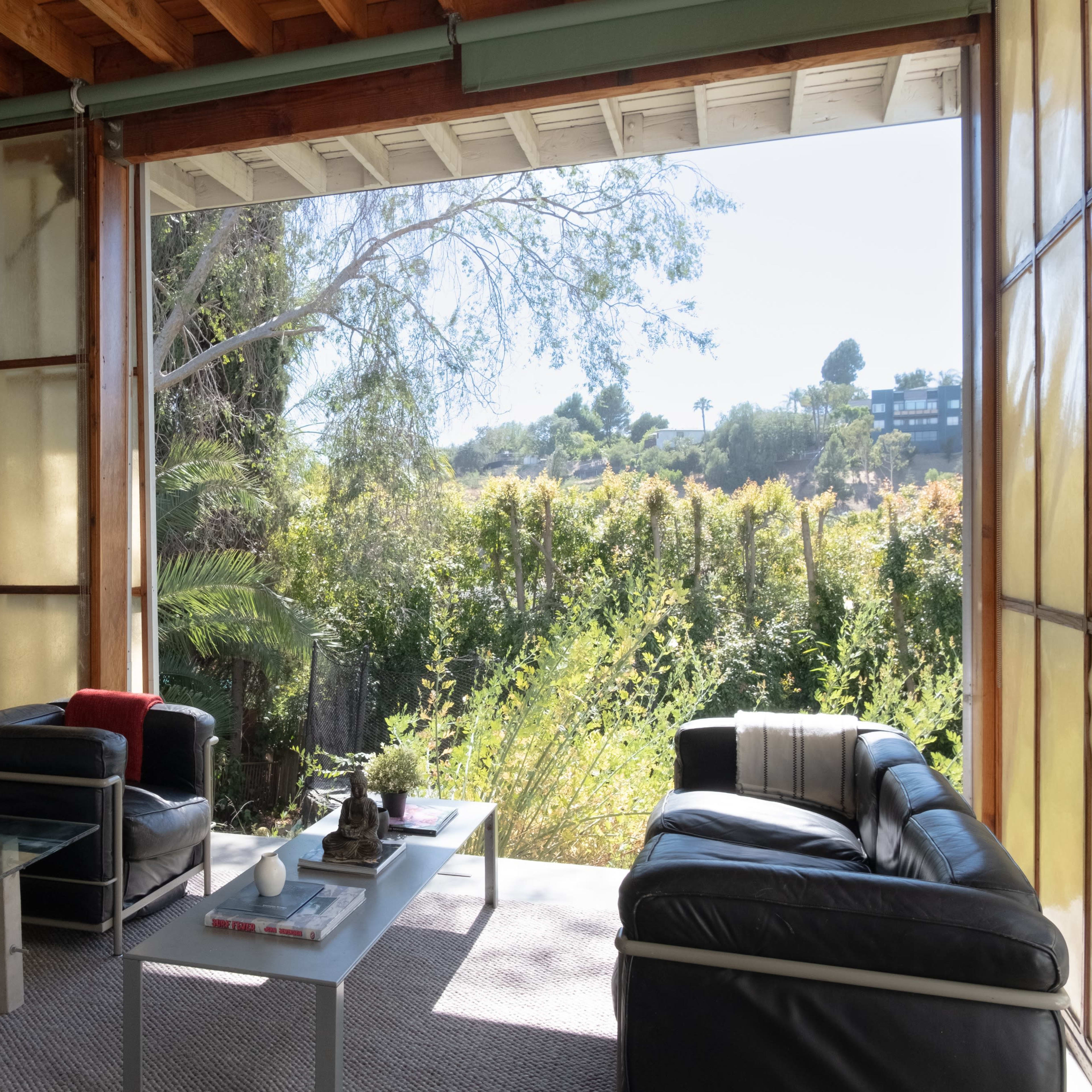 A modern living room features two sofas and a coffee table, with large sliding glass doors open to a lush outdoor garden and a clear sky view.