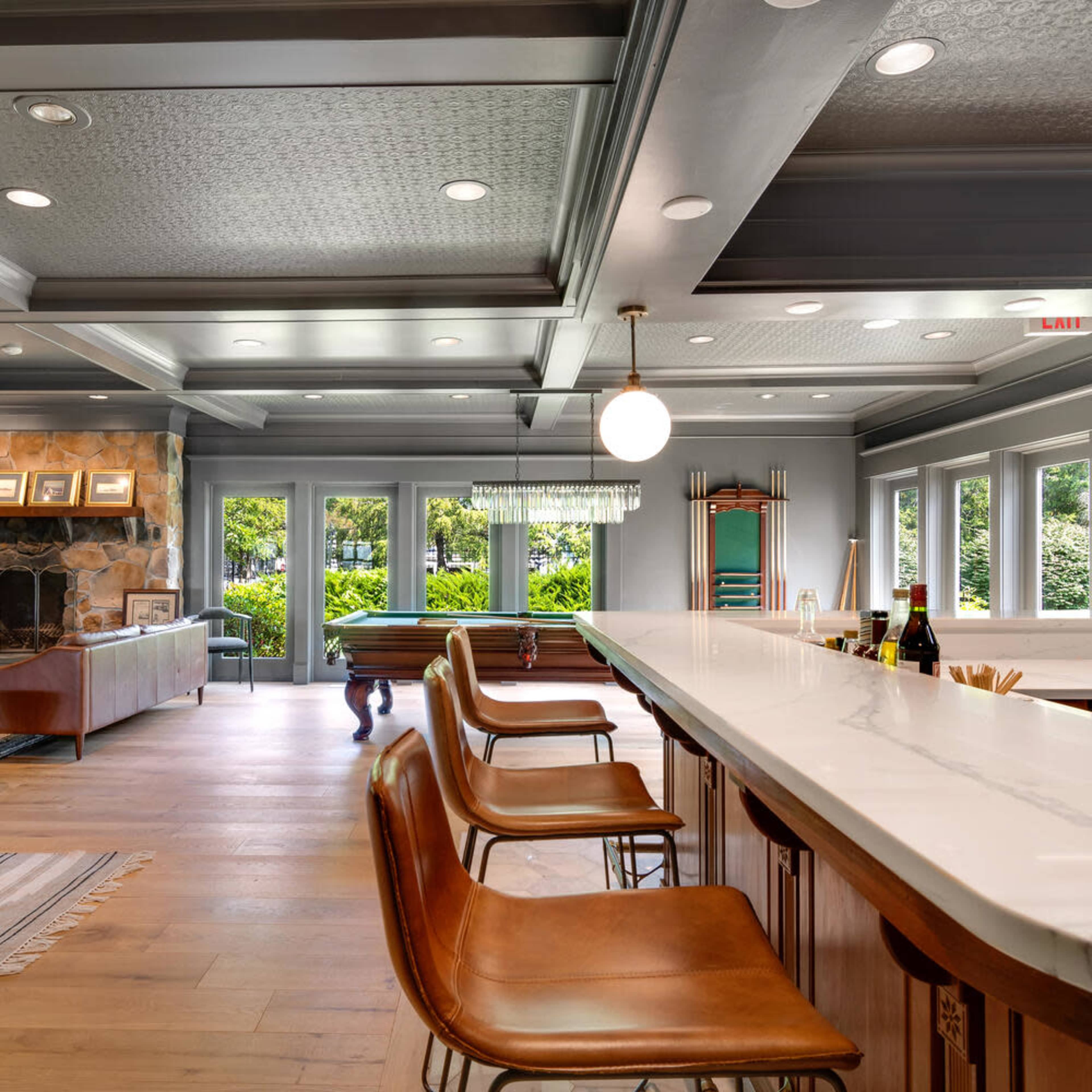 A spacious interior living area with a kitchen bar featuring four brown stools, a stone fireplace, and large windows overlooking greenery.