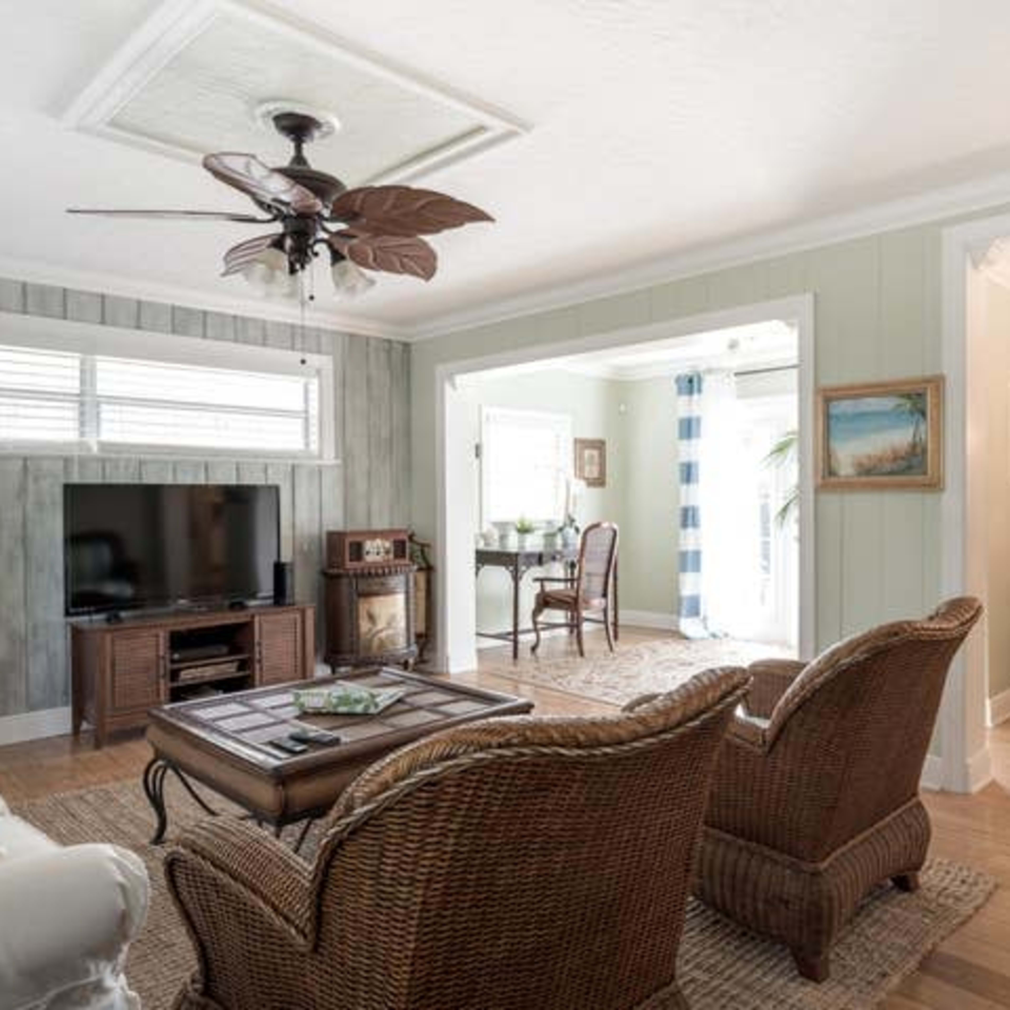 A well-lit living room with two wicker chairs facing a coffee table and a television, leading to an adjacent dining area with a small table and chairs.