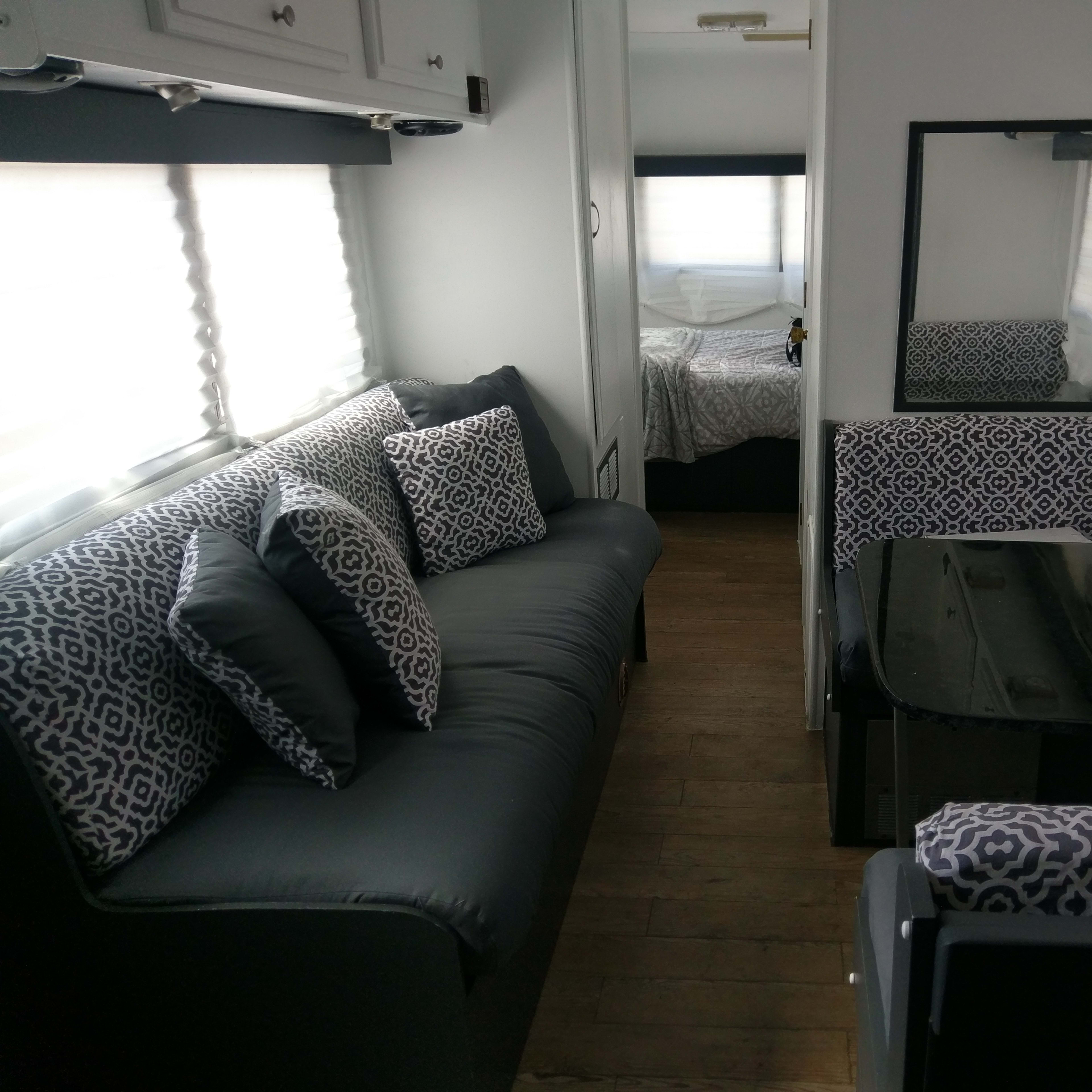 The interior of a camper trailer featuring a gray couch, patterned cushions, a black table, and a mirrored wall, with a bed visible in the rear.