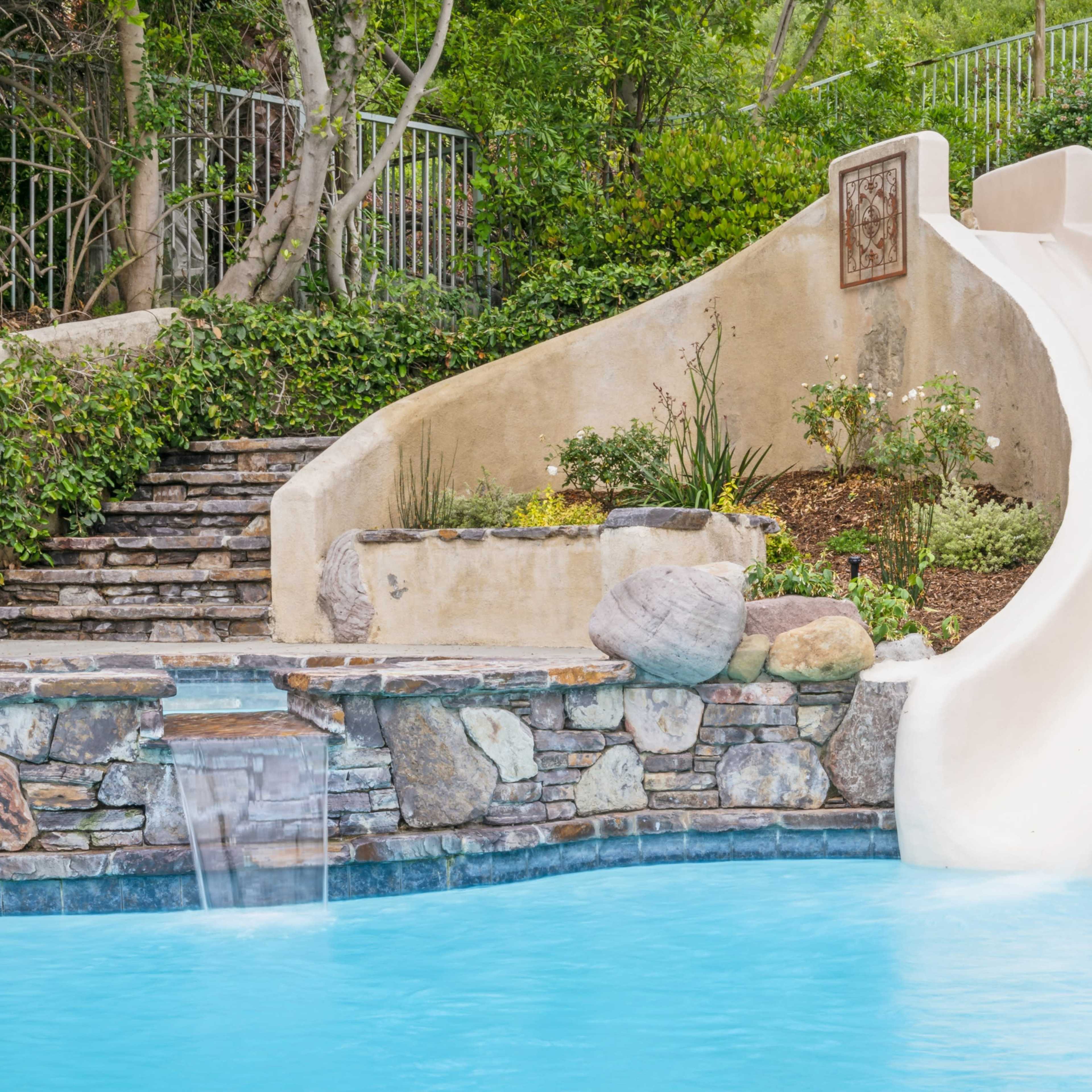 A white waterslide curves down into a blue swimming pool surrounded by stonework and greenery.
