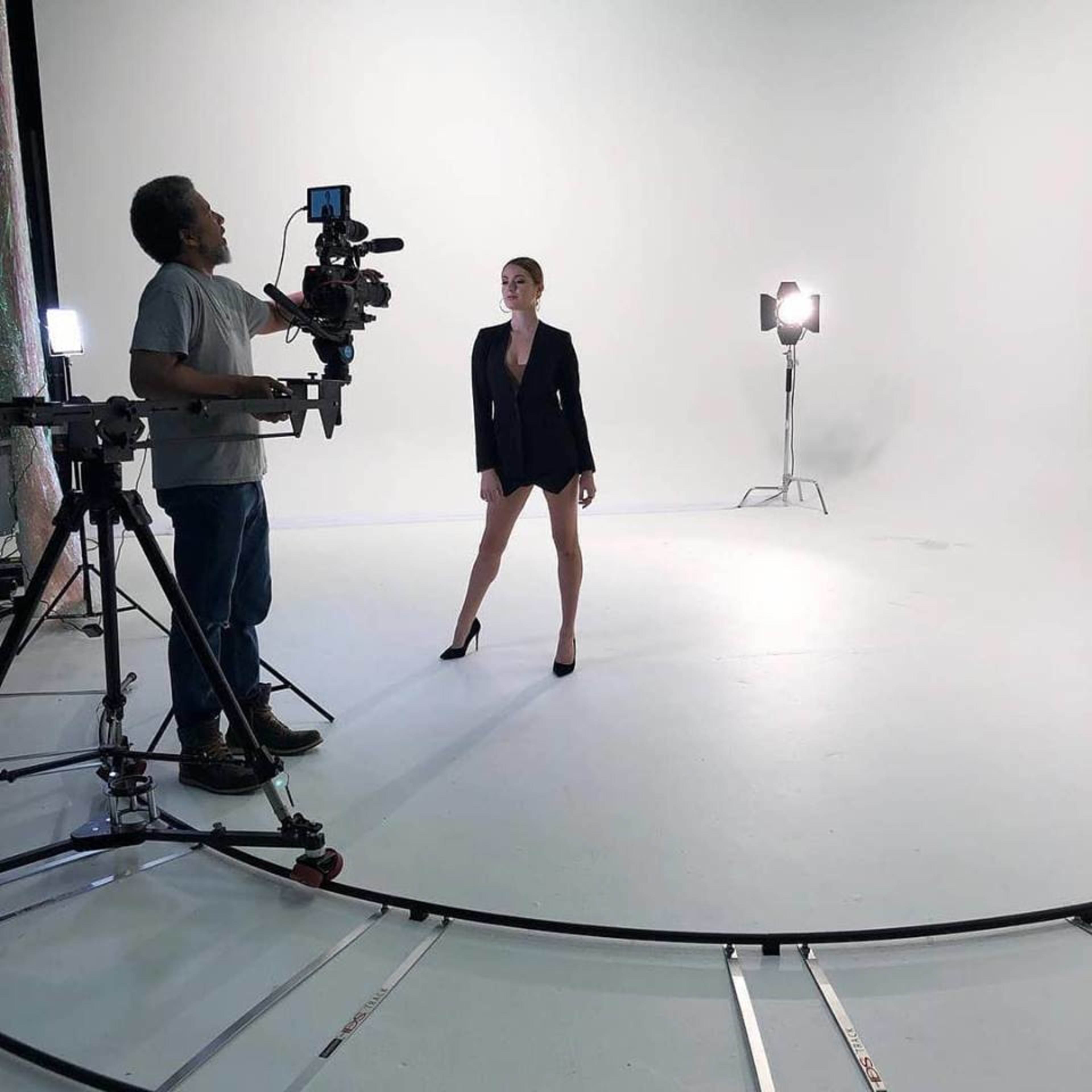 A person in a black blazer and high heels poses in a well-lit studio while a camera operator adjusts equipment.